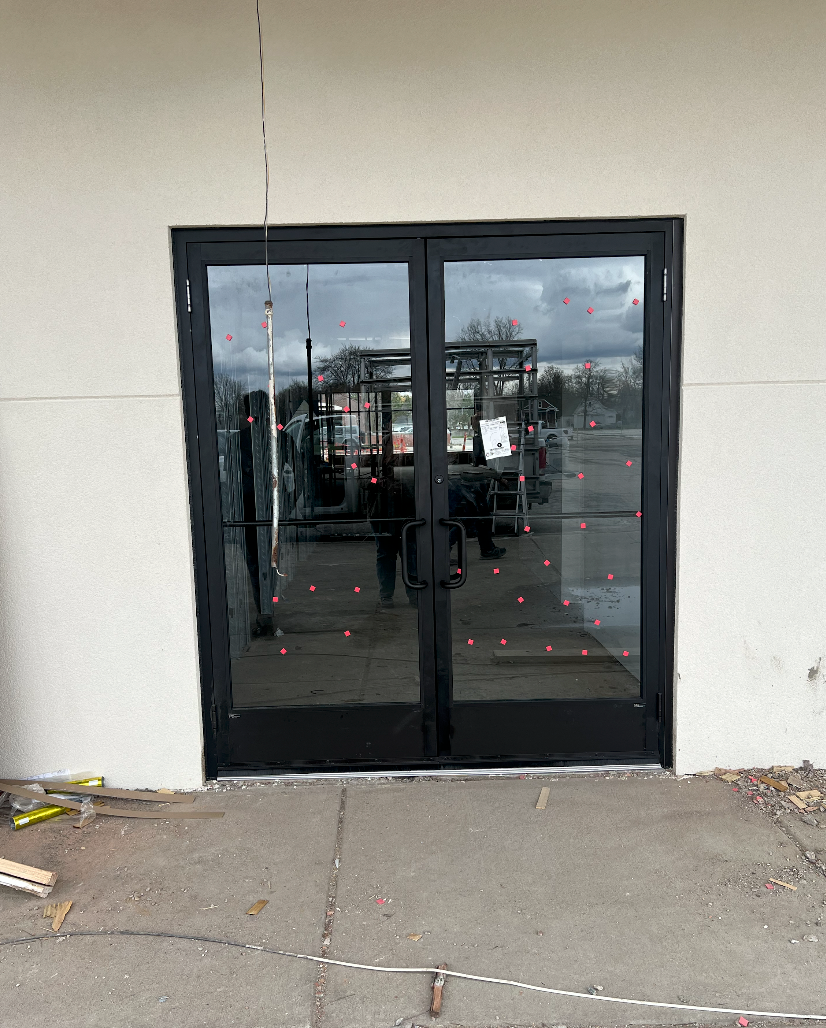 Black double doors with glass panels; storefront entrance. Concrete walkway, beige wall.