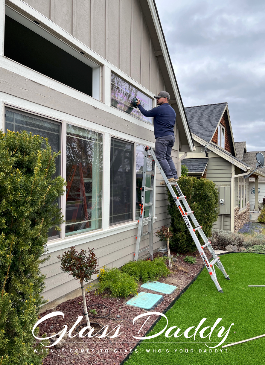 Man on ladder cleaning windows of a house with green lawn, sky, and