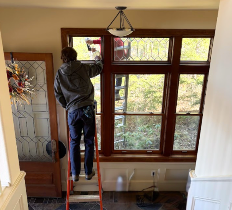 A person in a gray hoodie on a stepladder cleans the interior of large, multi-paned windows in a home foyer.