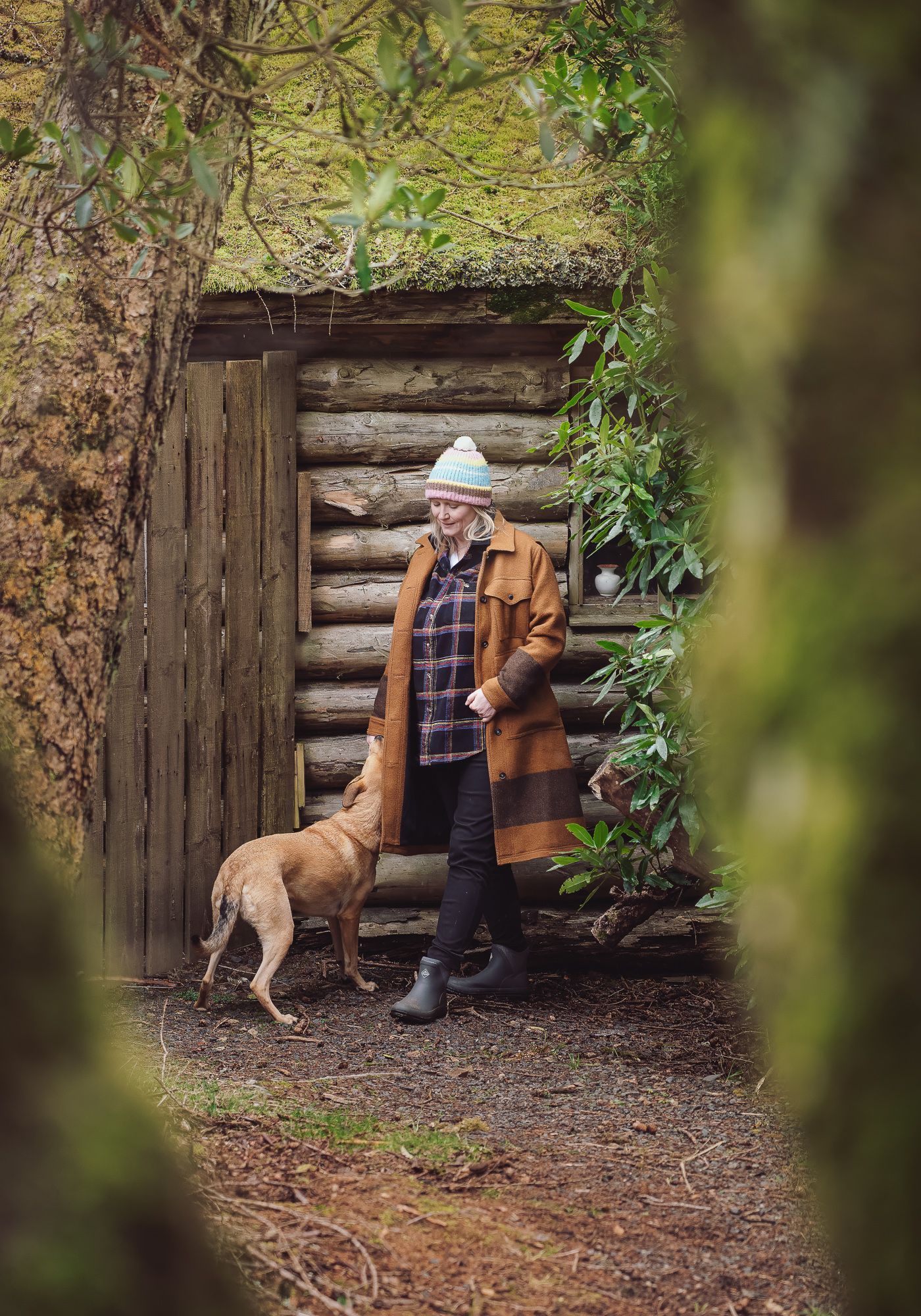 woman wearing long brown coat, standing with yellow dog in front of a log cabin