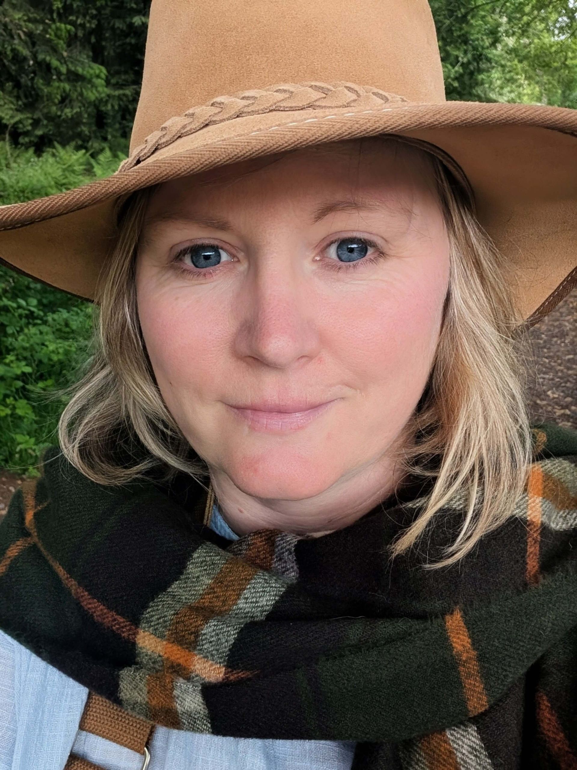 Claire Gibb from Lullabrae wearing a hat and plaid scarf smiles outdoors. Forest in the background.