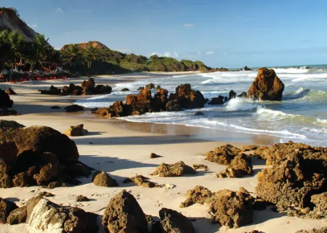 Praia rochosa ensolarada com ondas, faixa de areia e falésias verdes ao longe.
