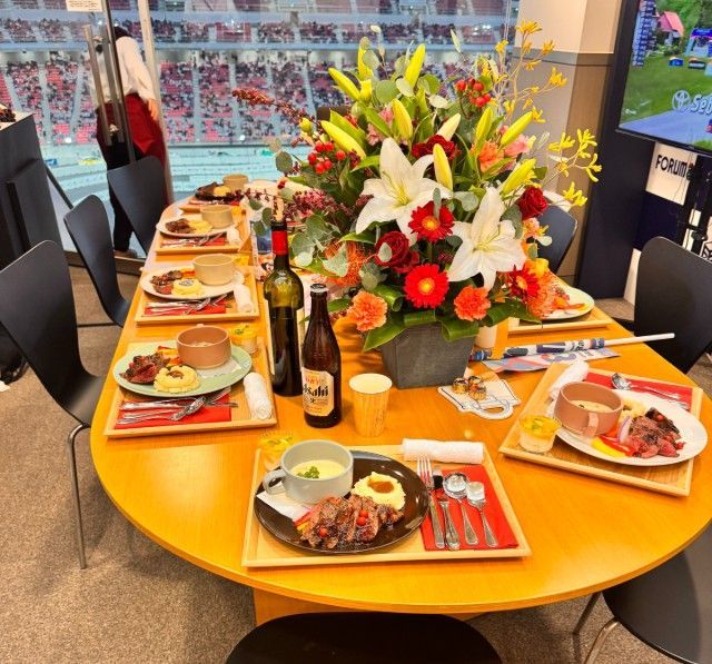 食事用に皿、飲み物、フラワーアレンジメントが置かれた楕円形のテーブル。背景にはスタジアムの景色が見える。