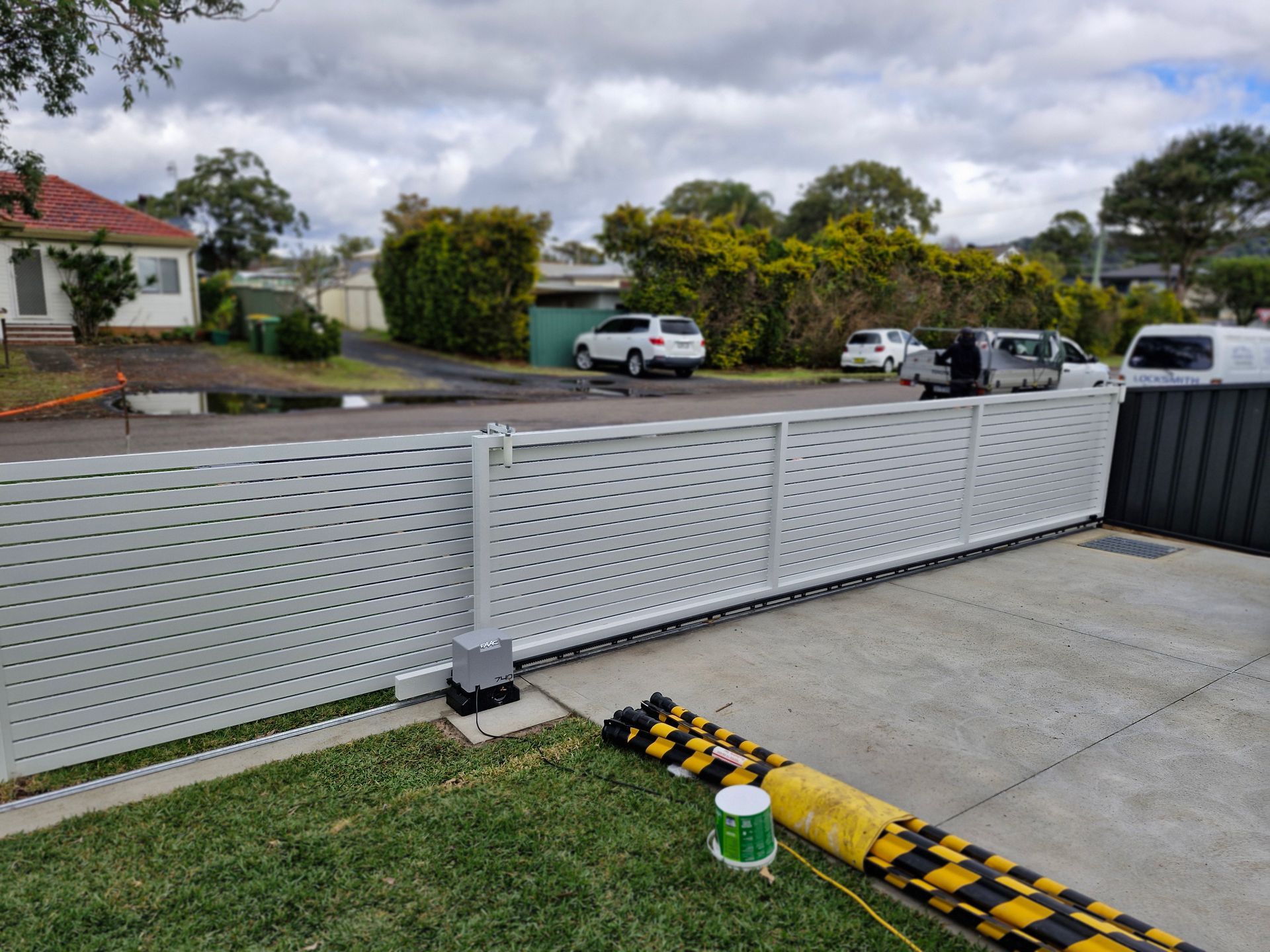 Gray, automated sliding gate across driveway, with vehicles and houses in the background. — Strike Fencing & Gates In Tuggerah, NSW