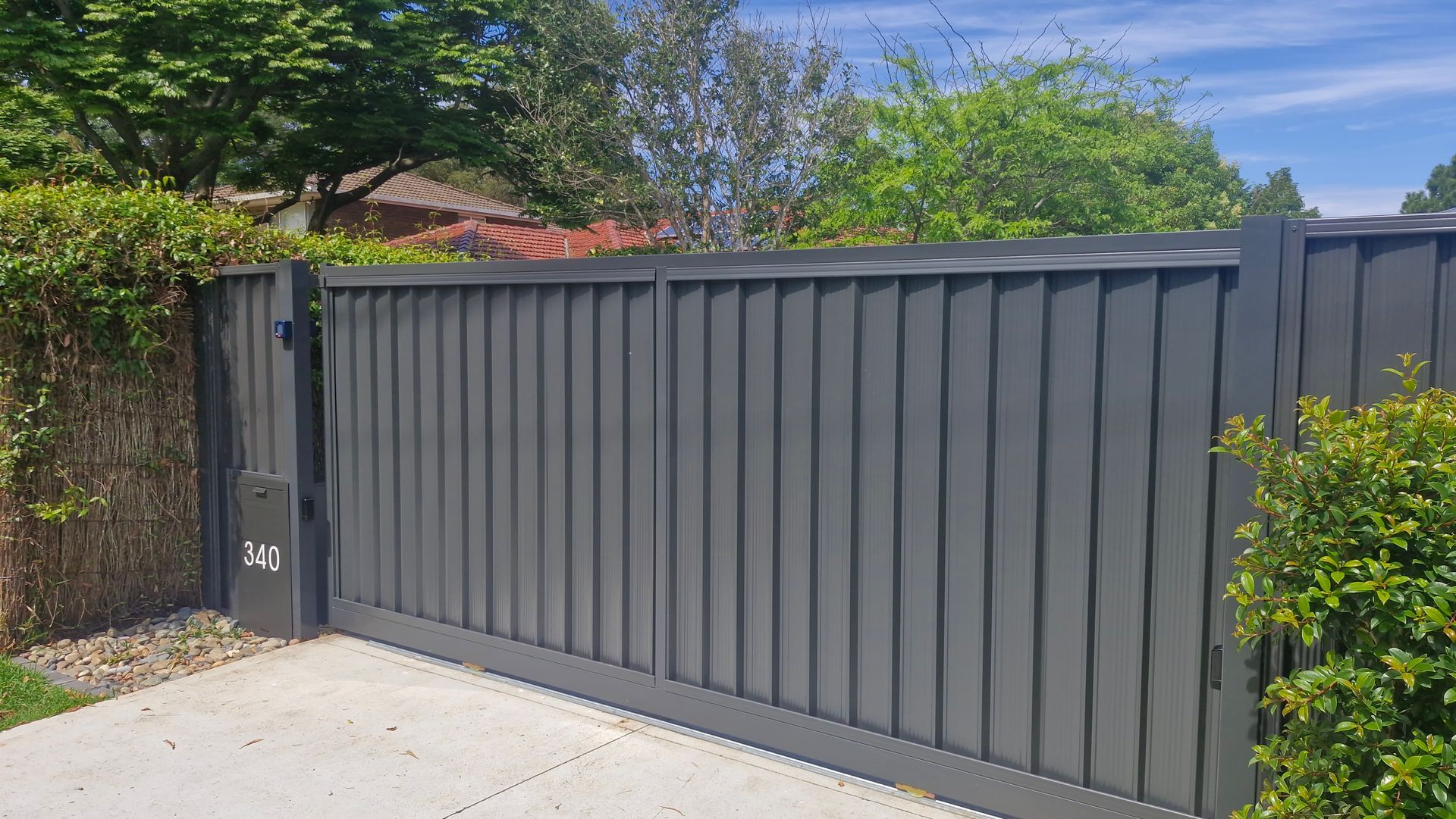 Gray metal driveway gate with vertical slats, set against green foliage.