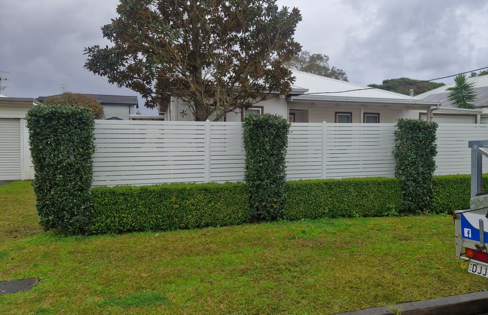White slat fence with hedges, green grass, and a house in the background on an overcast day. — Strike Fencing & Gates In Tuggerah, NSW