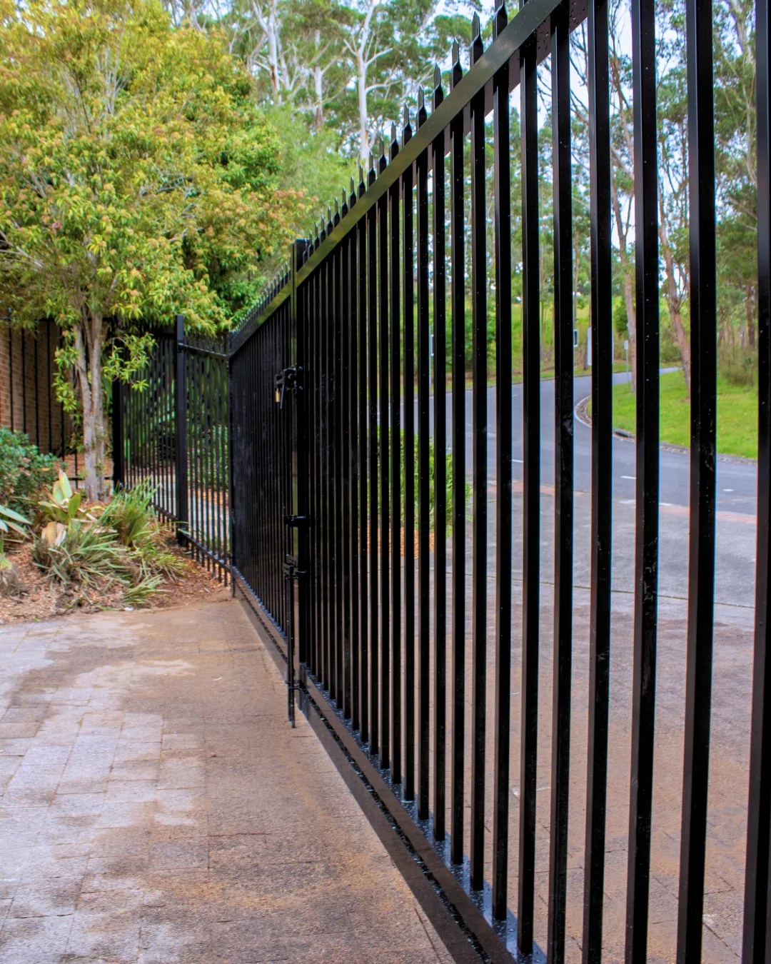 Black Metal Security Fence With Vertical Bars Beside a Paved Path and Driveway — Strike Fencing & Gates In Tuggerah, NSW