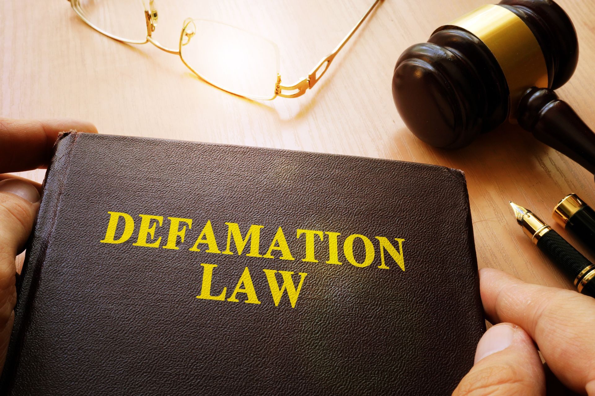 Hands holding a book titled “DEFAMATION LAW” beside a judge’s gavel and eyeglasses