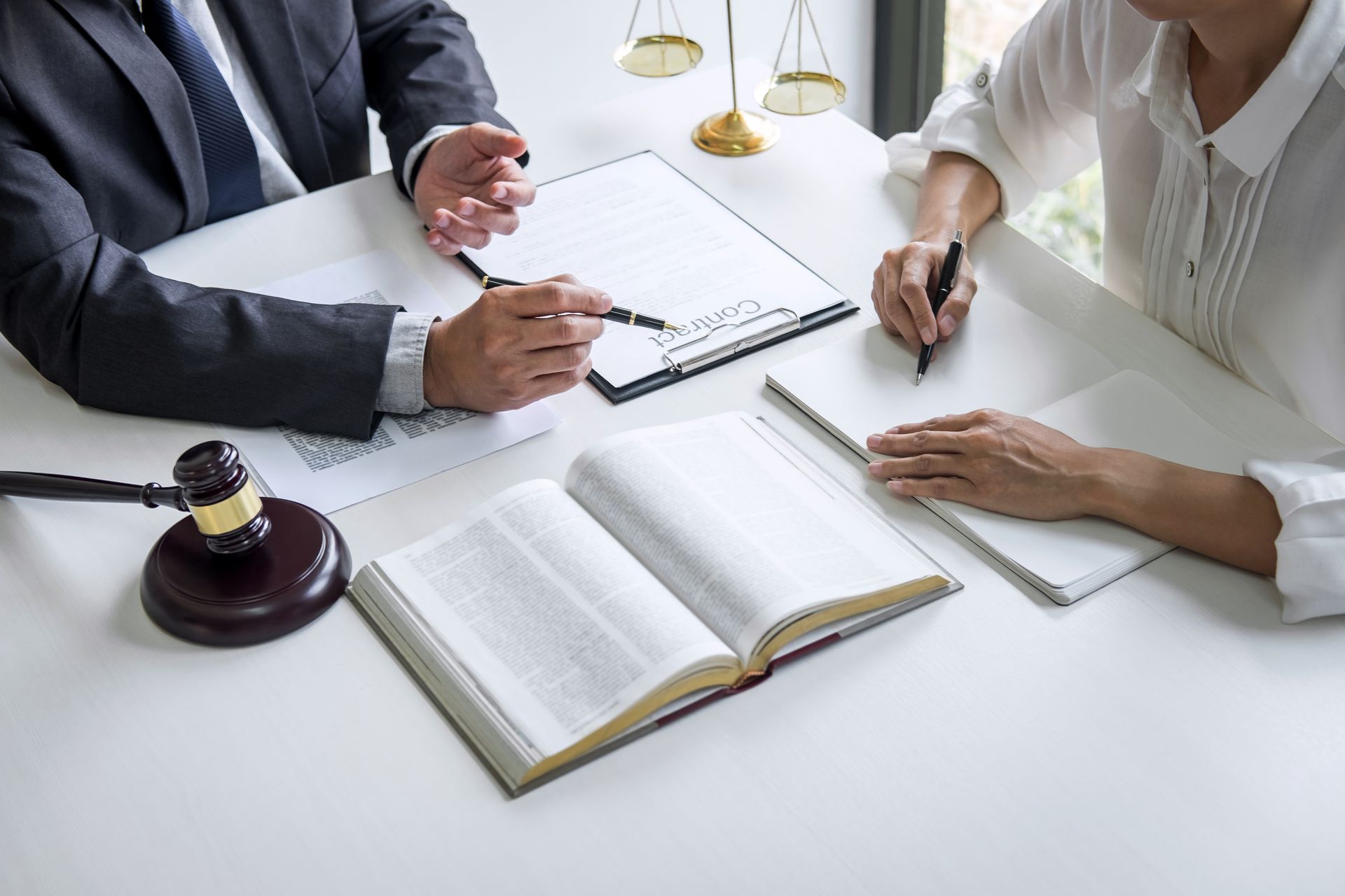 A professional in a suit and a person in a white shirt sit at a desk with a law book, a gavel, and legal documents.