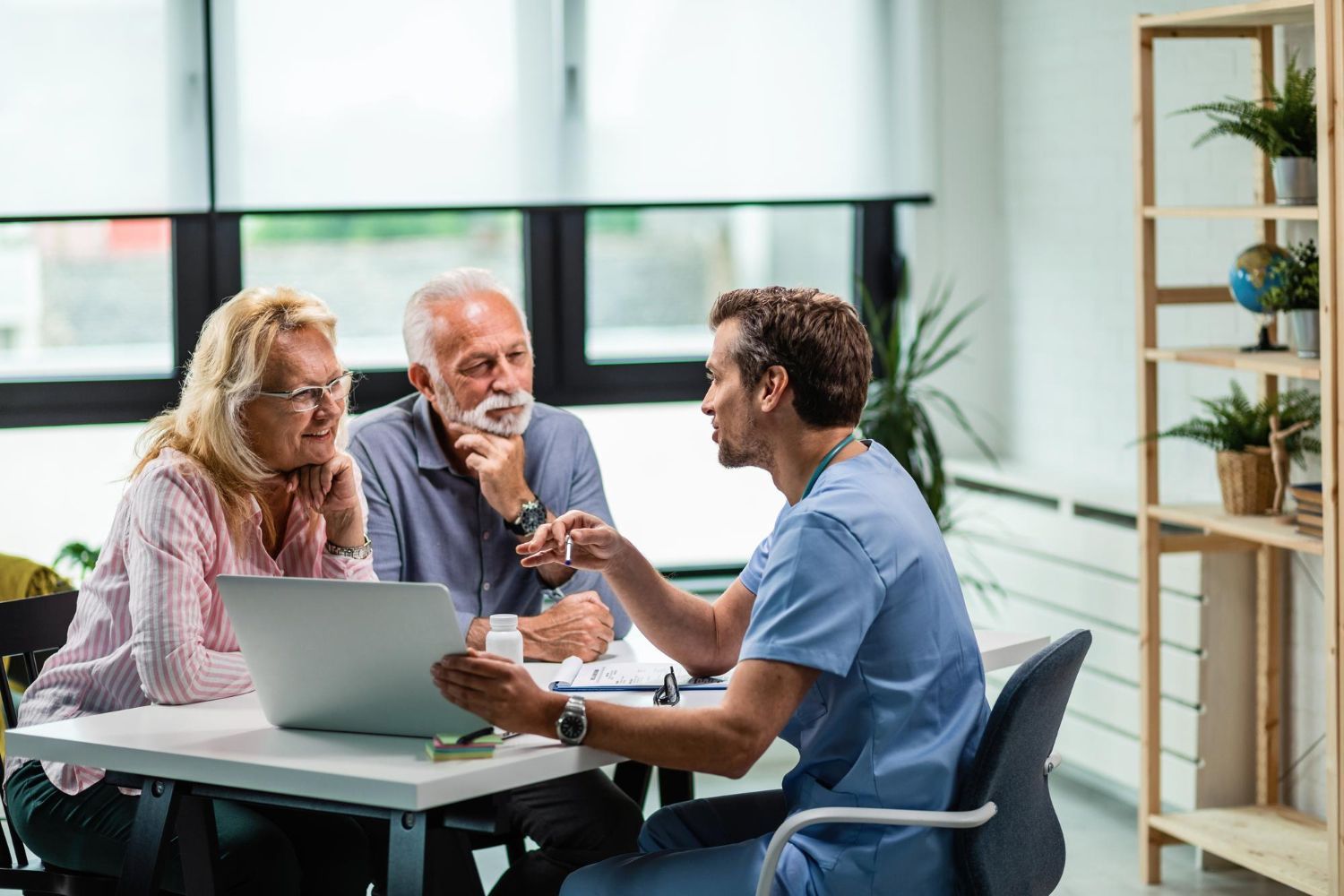 A doctor in scrubs showing a couple information on a laptop in a brightly lit office.