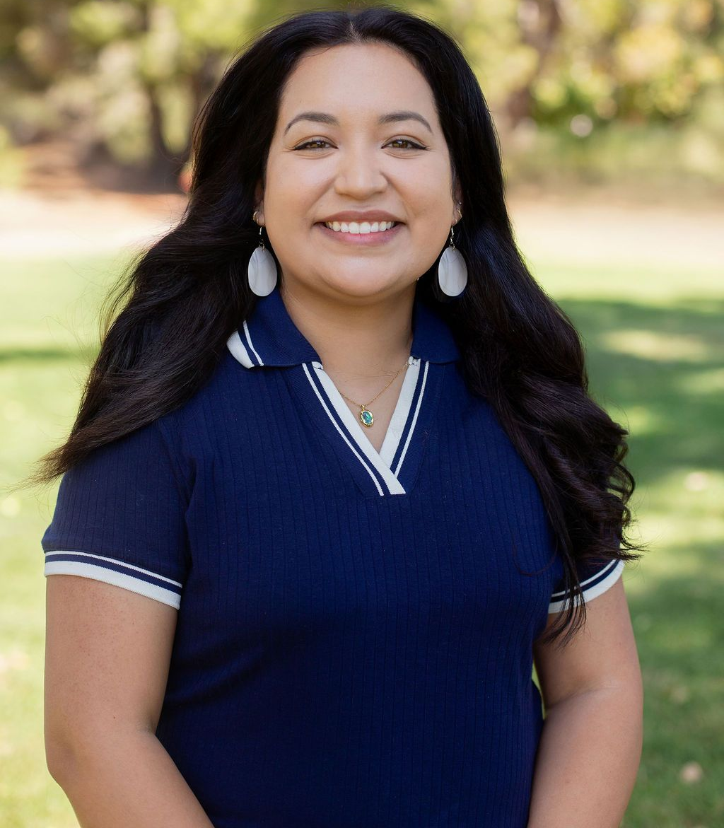 Woman with dark hair smiles, wearing blue striped shirt and white earrings, outdoors in sunlight.