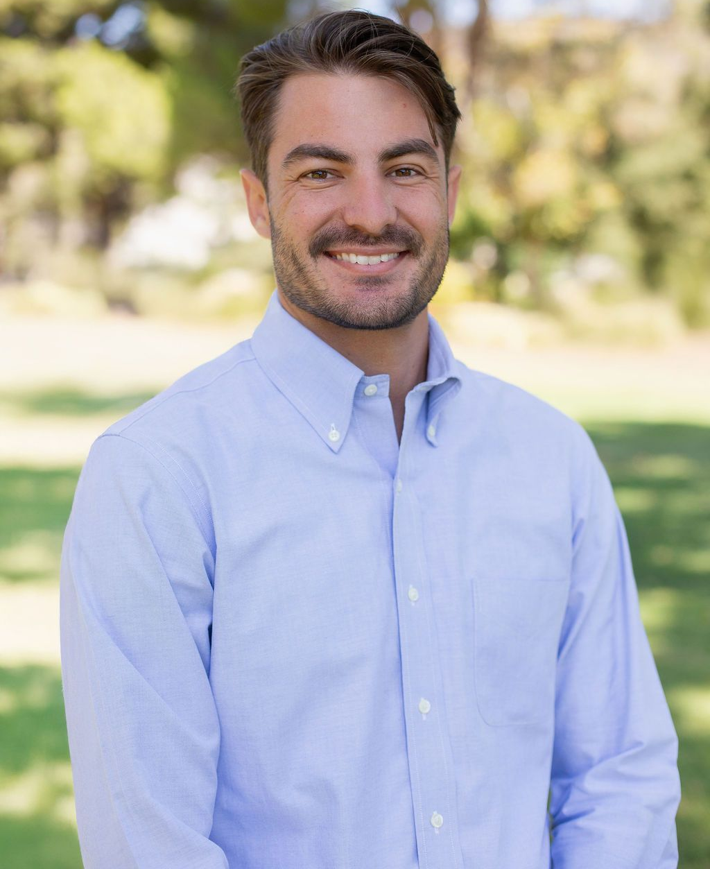 Man in a light blue button-down shirt smiles in an outdoor setting with trees in the background.