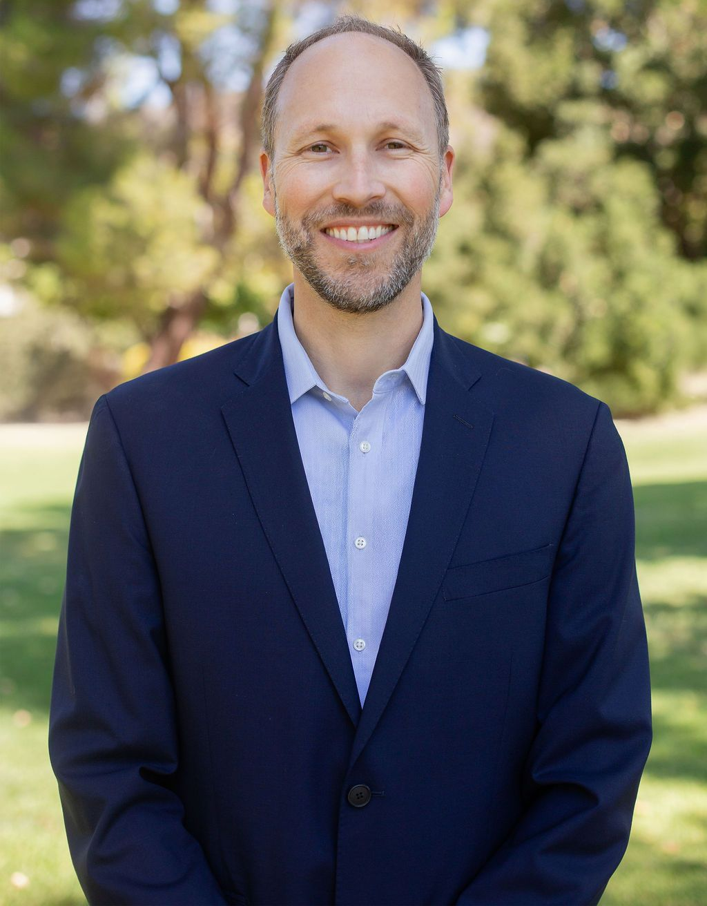 Man in a blue blazer and shirt smiling outdoors in front of trees.