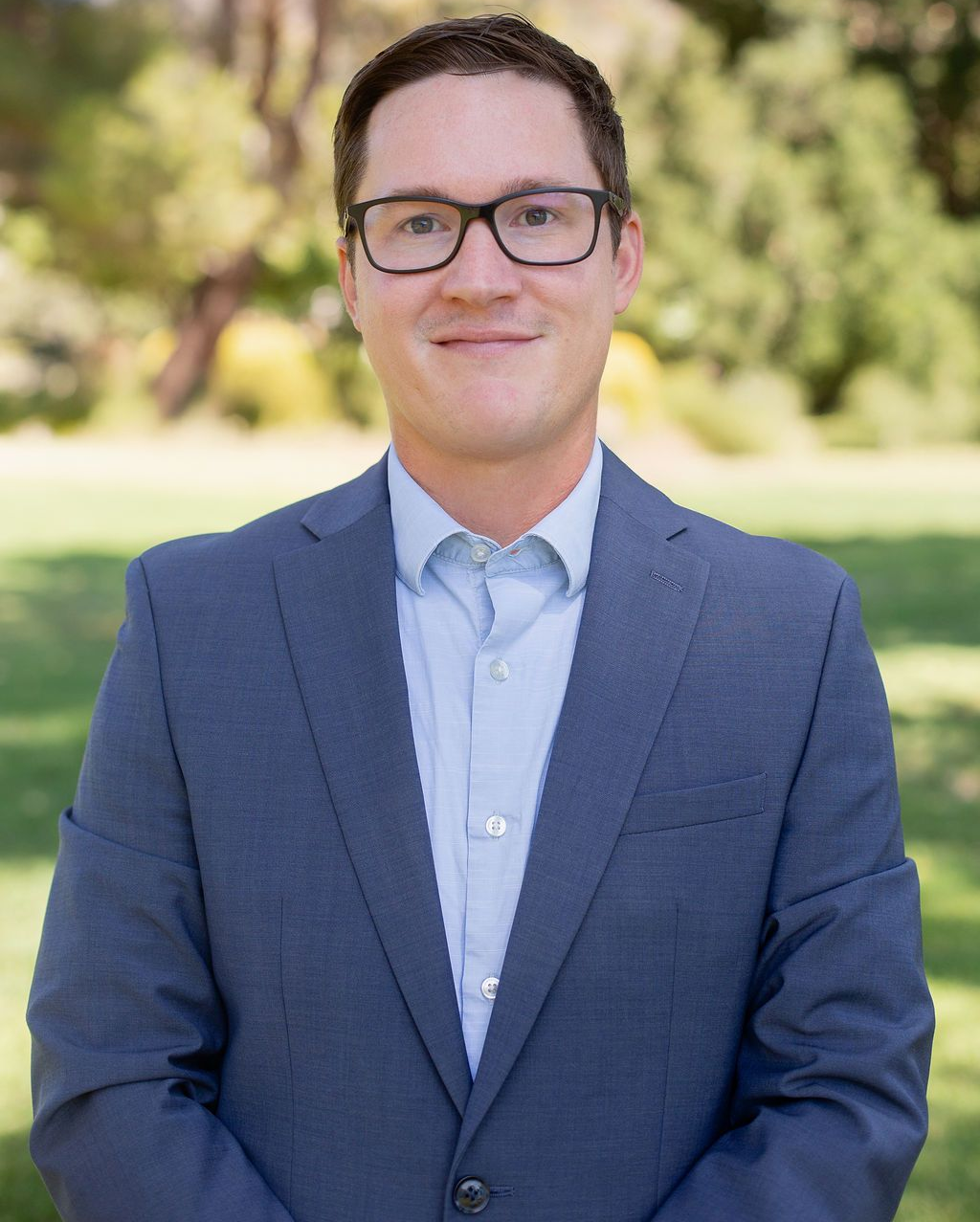 Man wearing glasses and a blue suit, standing outdoors, smiling.