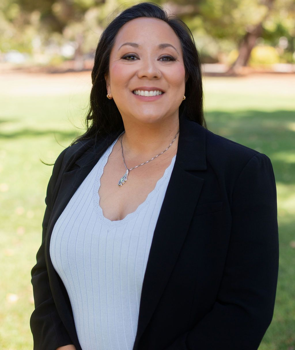 Woman in black blazer, smiling, outdoors in front of greenery.