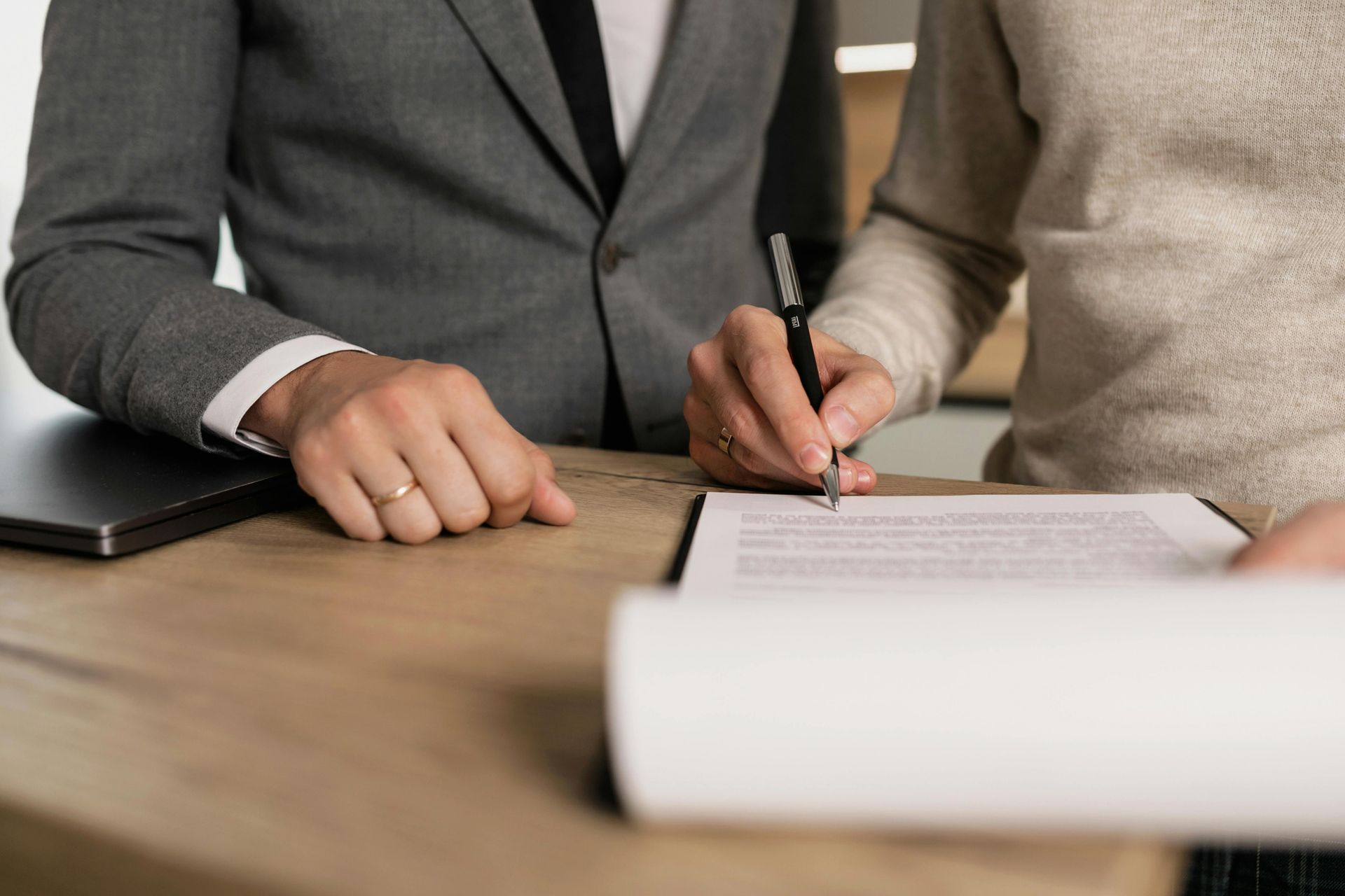 Person in a suit signing a document with another person. A laptop sits on the table.