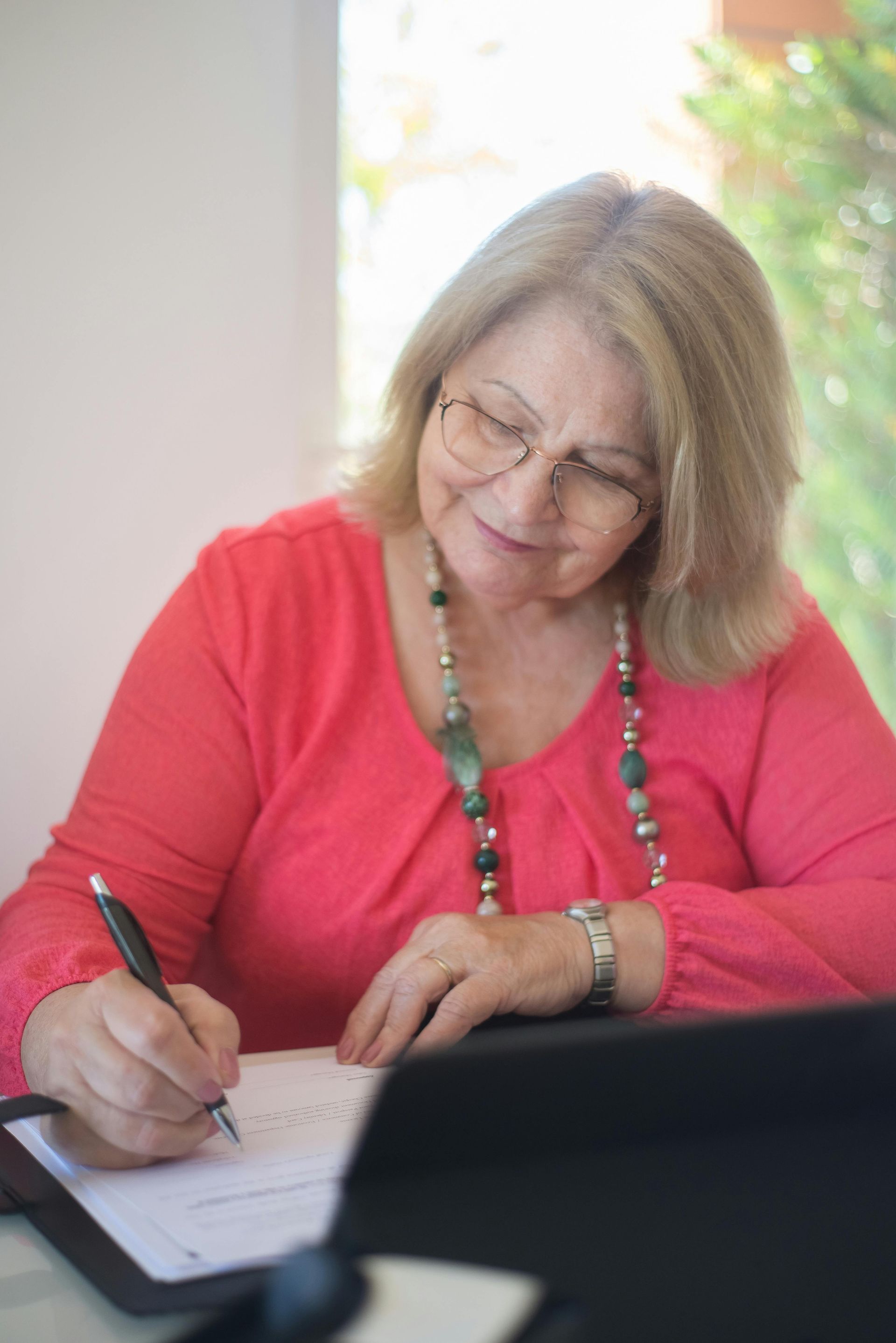 Woman in coral top writing on paper, wearing glasses and a beaded necklace.