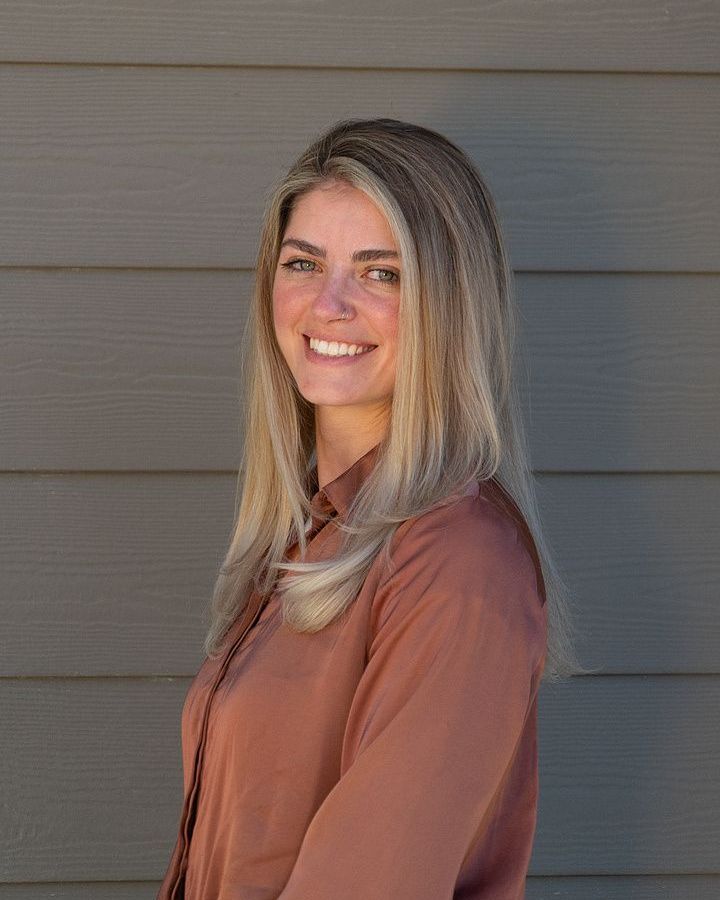 A woman in a brown shirt is smiling in front of a gray wall.