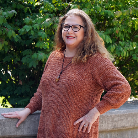 A woman wearing glasses and a sweater is standing in front of a tree.