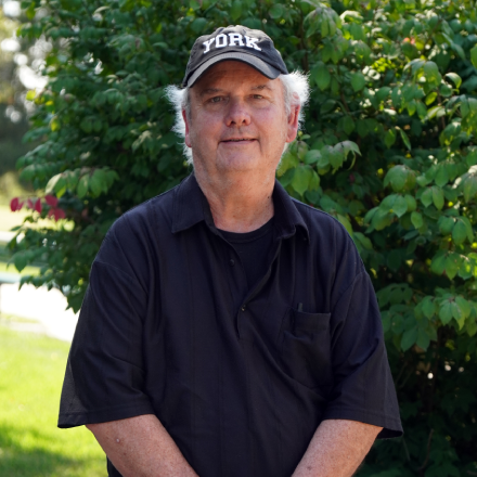 A man wearing a york hat stands in front of a bush