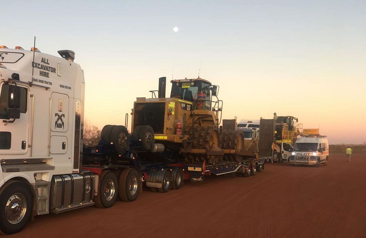 A white semi truck is carrying a bulldozer on a dirt road.