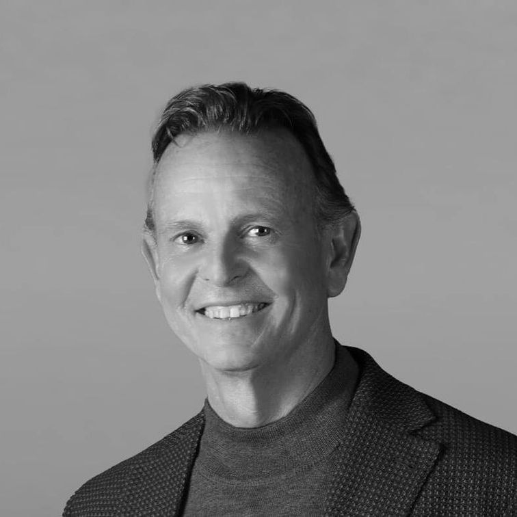 A man in a suit and sweater is smiling in a black and white photo.