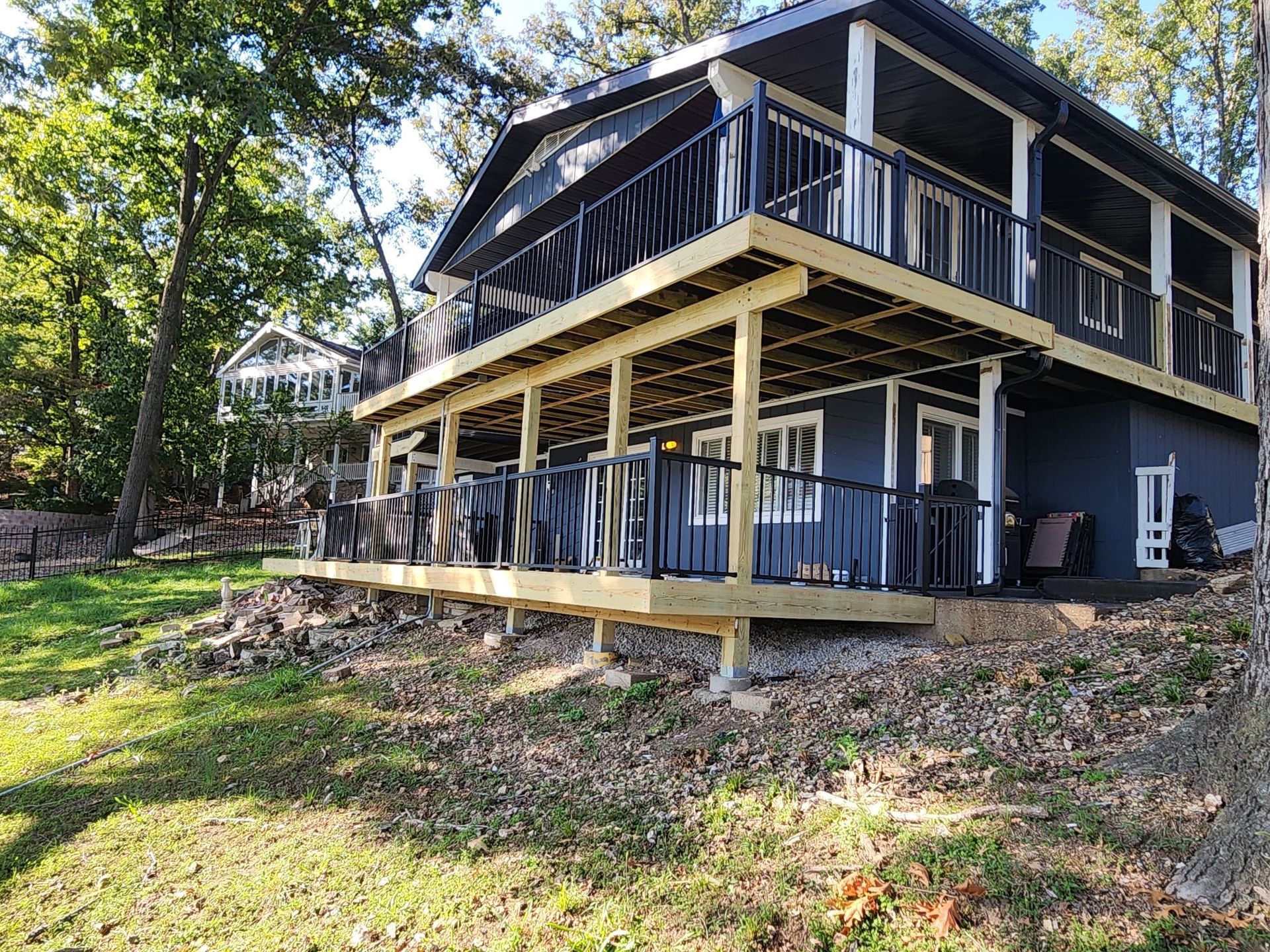 A two-story house with dark gray siding and a large, newly built light wood deck, nestled in a wooded, grassy landscape.