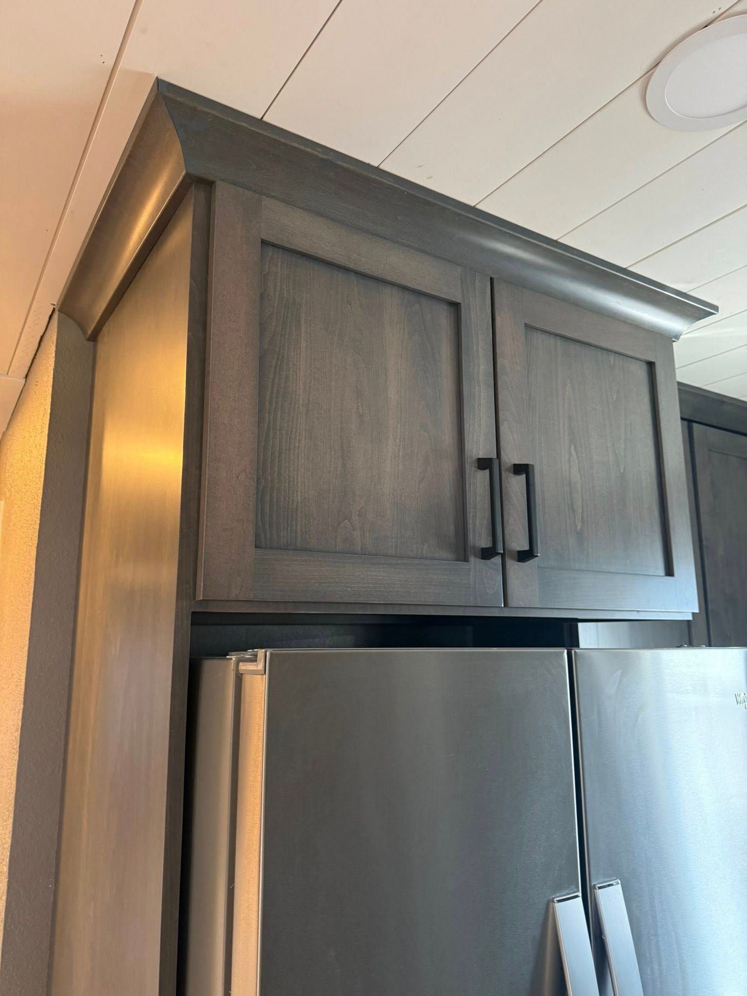 Dark grey kitchen cabinets installed above a stainless steel refrigerator, featuring crown molding and black handles.