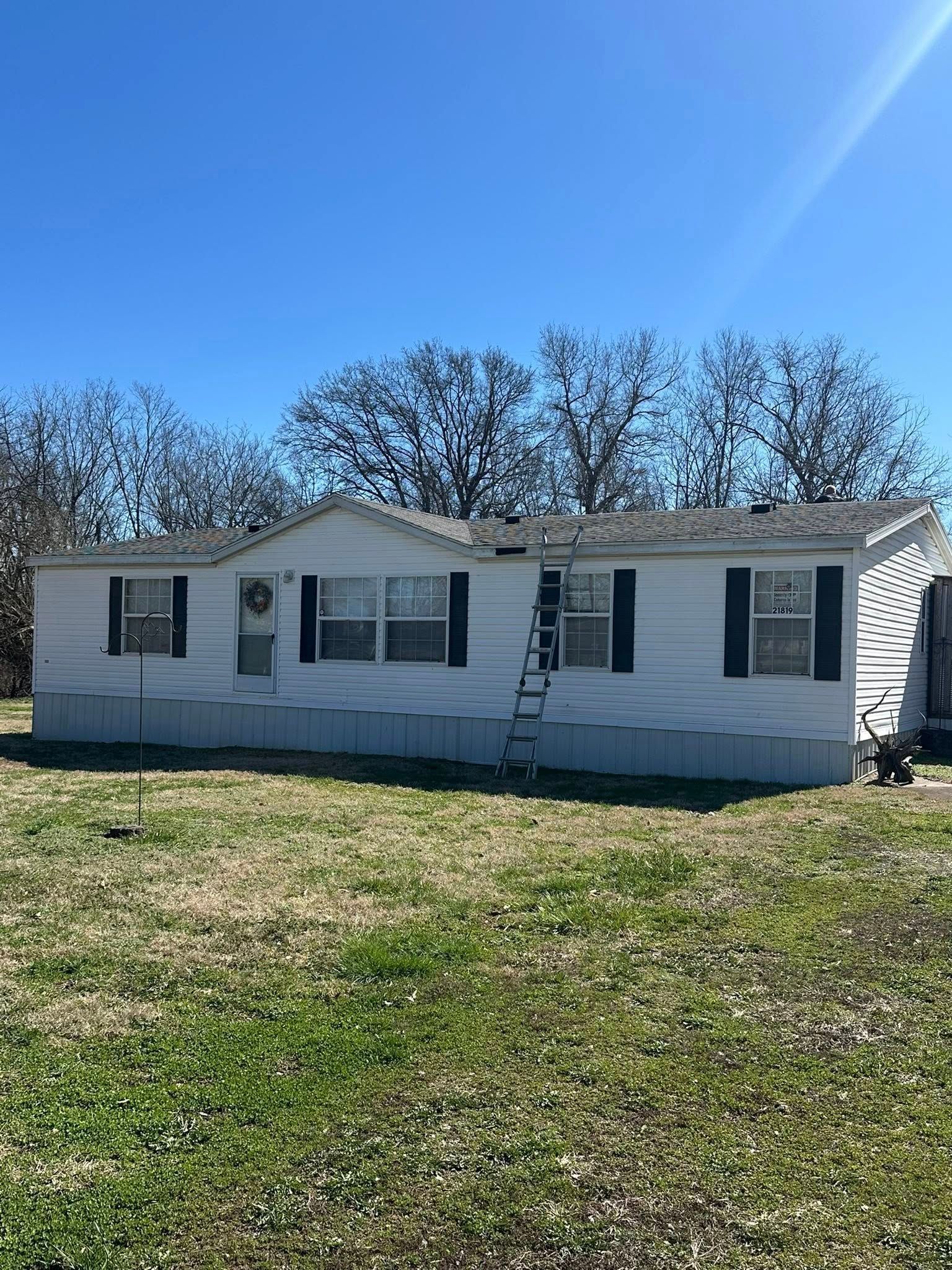 A white single-wide mobile home with dark shutters sits in a grass field under a clear blue sky.