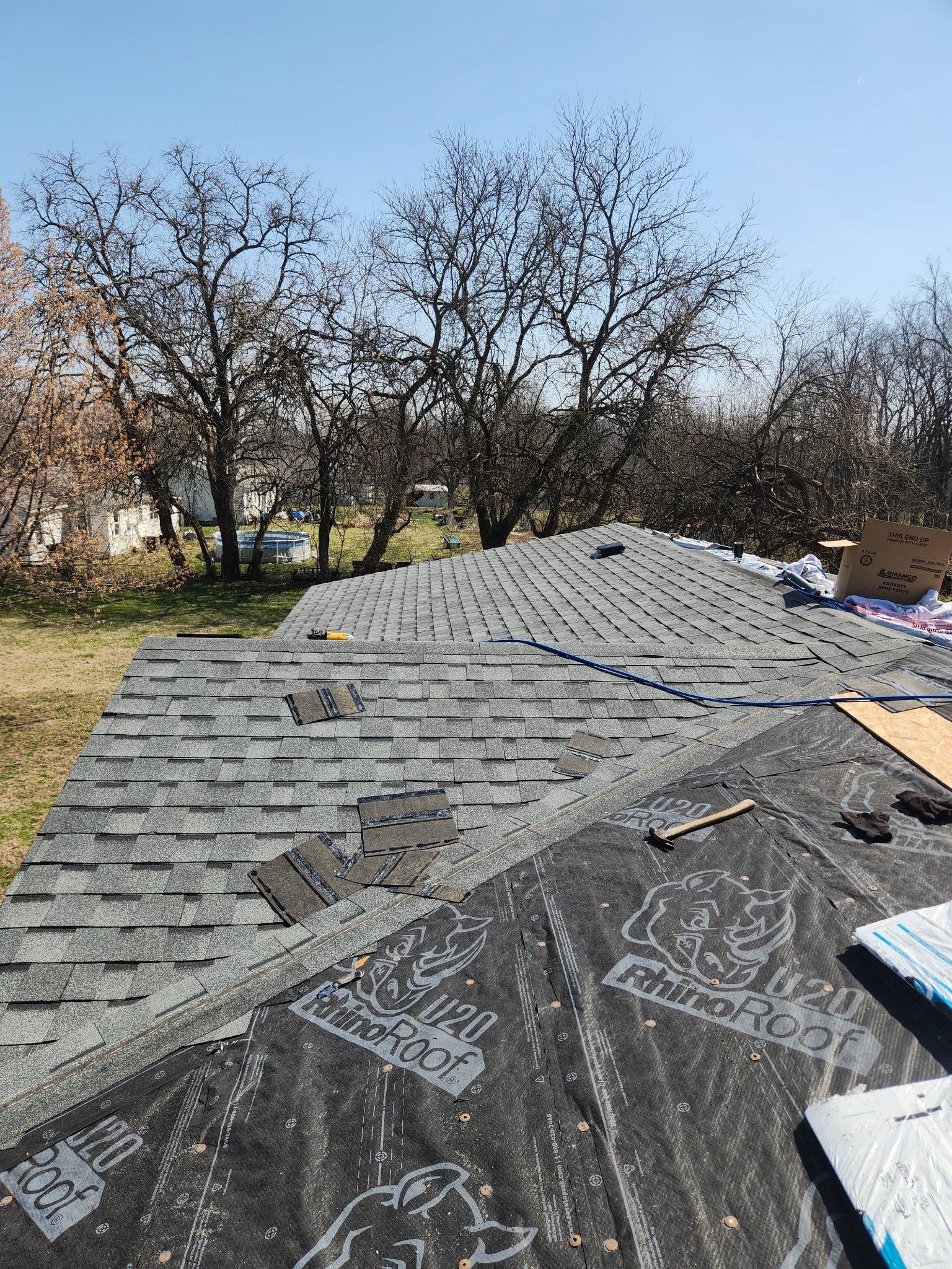 A roof under construction with partially installed gray shingles over black underlayment, set against bare trees and sky.