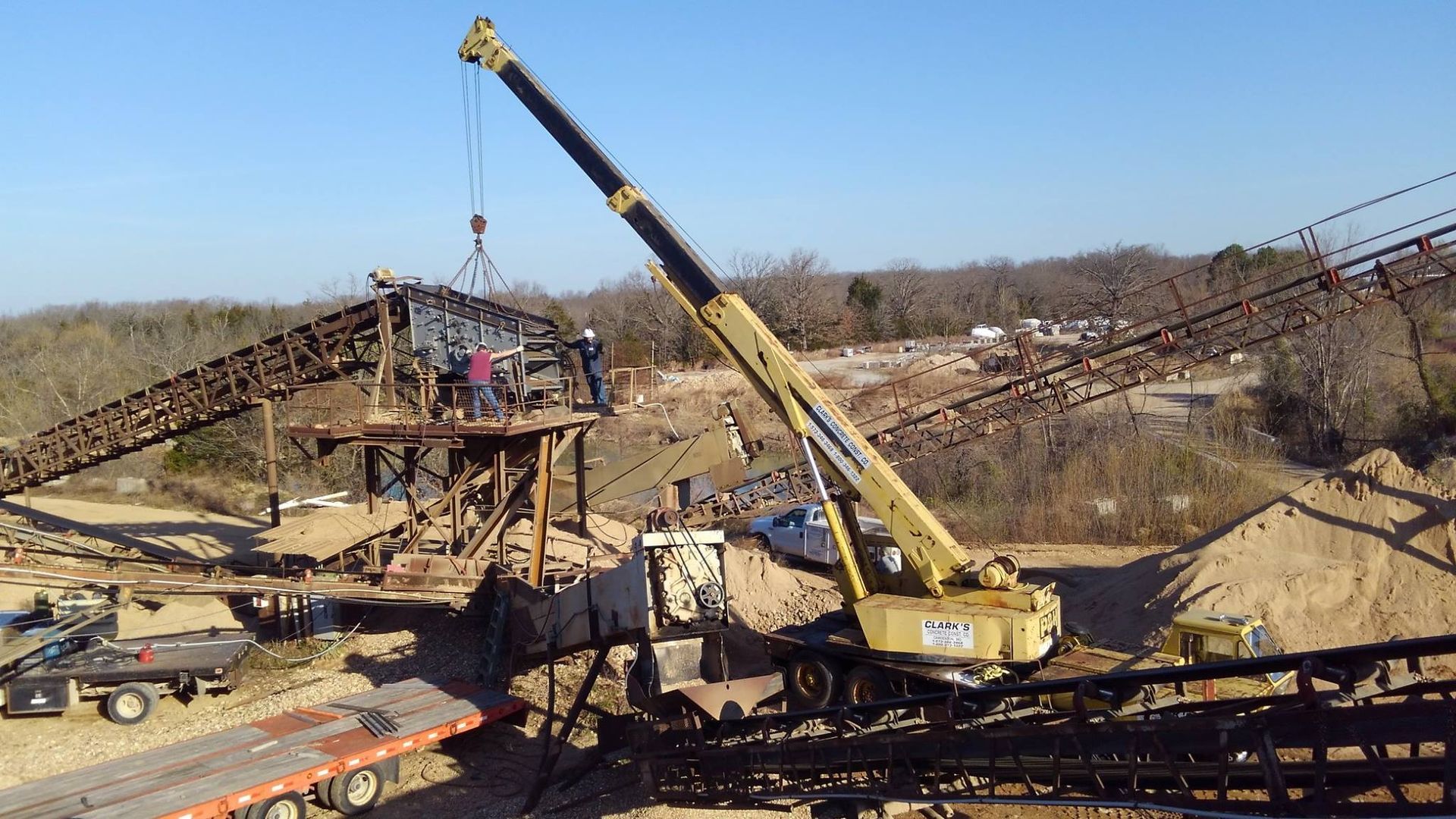 Crane lifting a structure at a construction site. Men work on the structure; sunny outdoor setting with trees and debris.