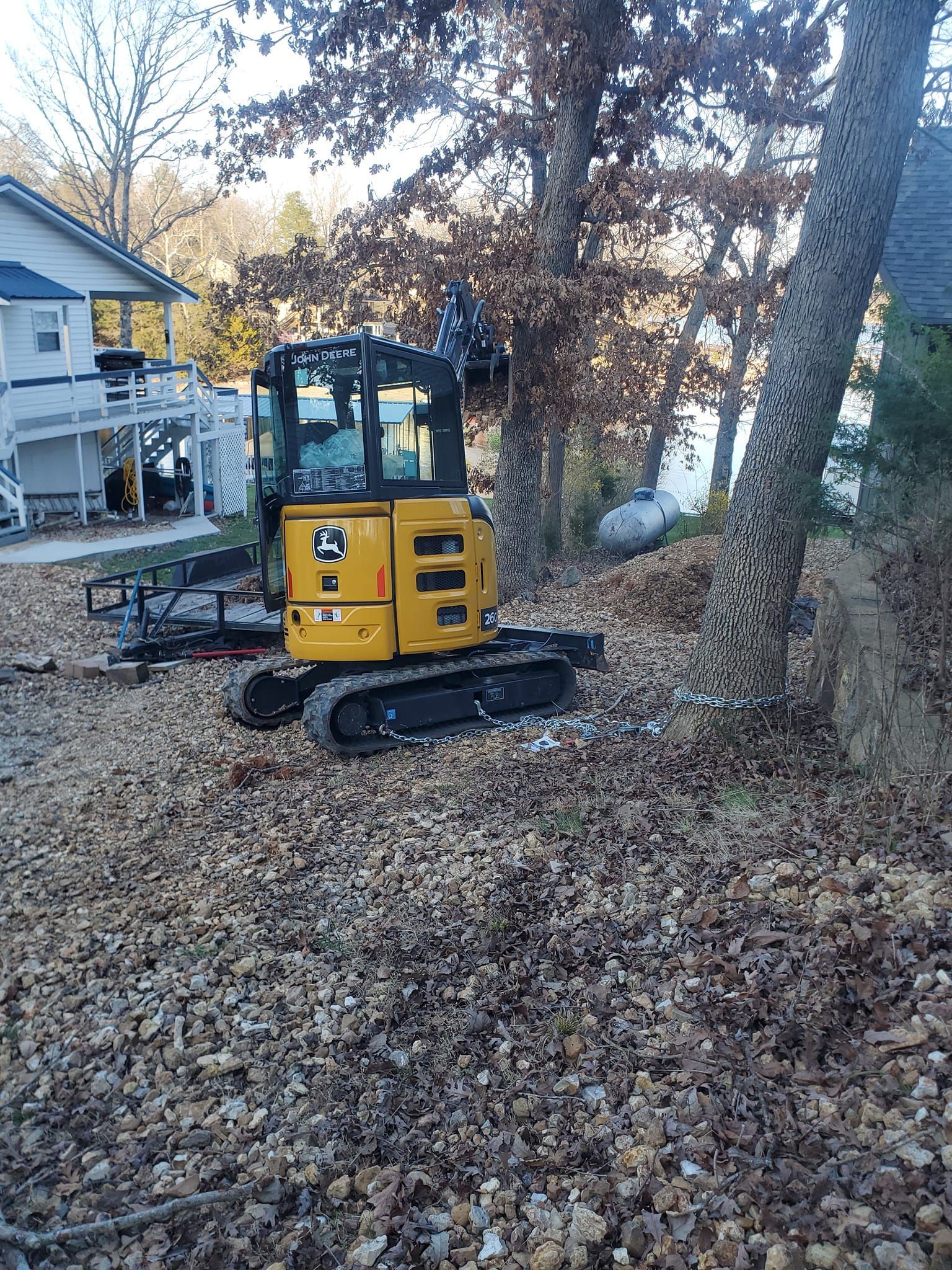 Yellow John Deere excavator on a leaf-covered shore near a house and trees.