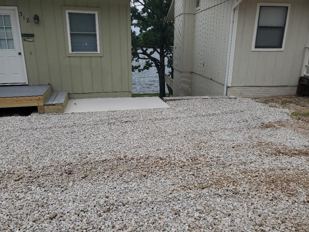 Gravel yard between two buildings, with a view of water and a tree in the background.
