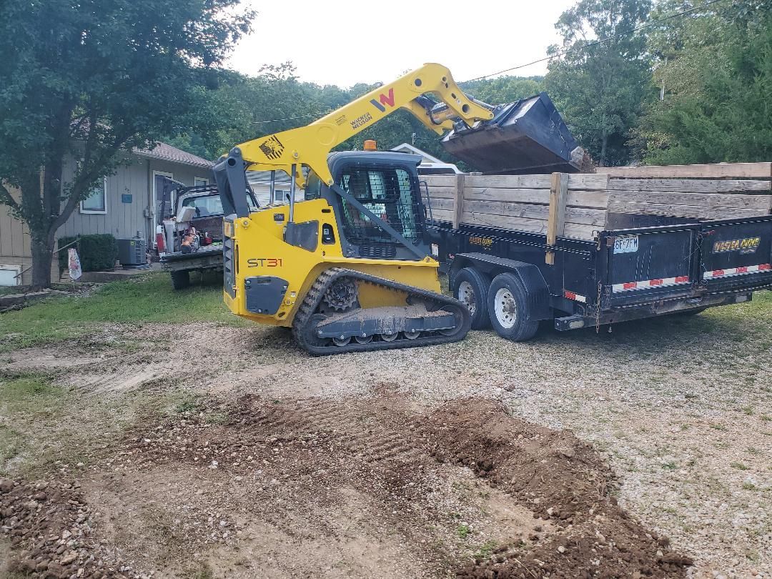 Yellow skid steer loading dirt into a black trailer on a gravel driveway.