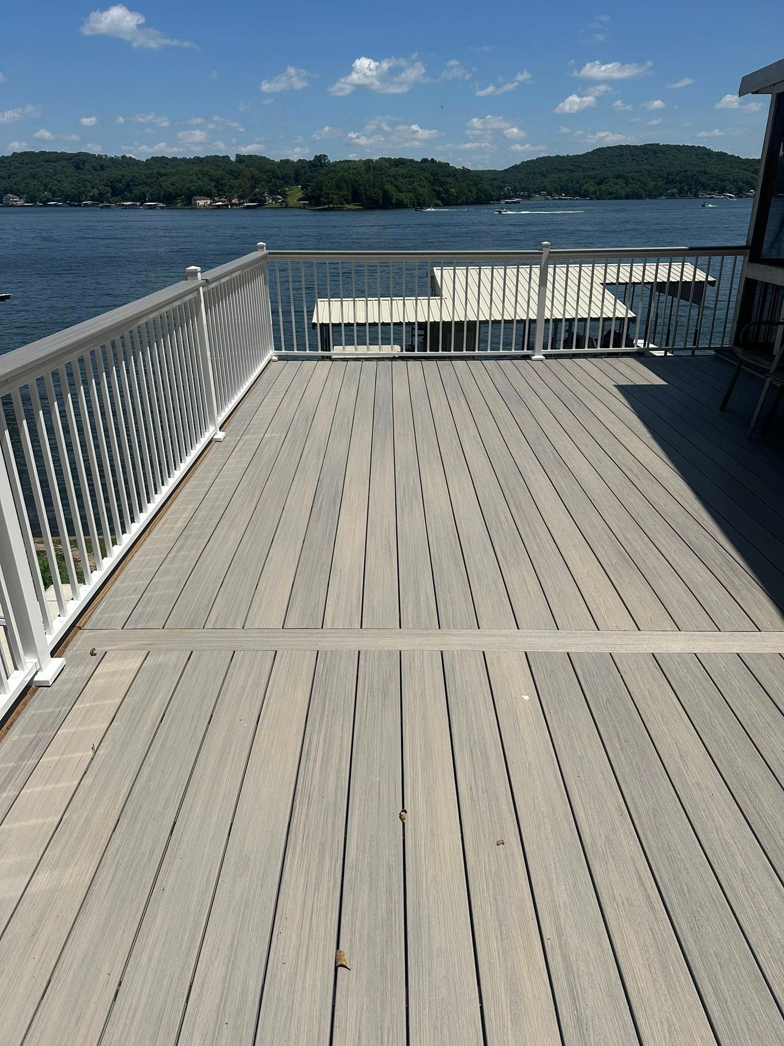 Deck overlooking a lake; white railing, wooden planks, and distant trees under a blue sky.