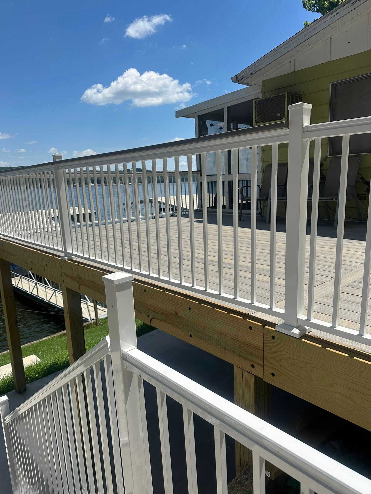 White railing surrounds a deck overlooking water, stairs lead down. Blue sky with clouds.