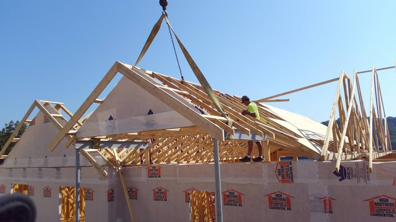 Crane lifting roof trusses onto a partially constructed house; a worker stands on the roof. Bright blue sky.
