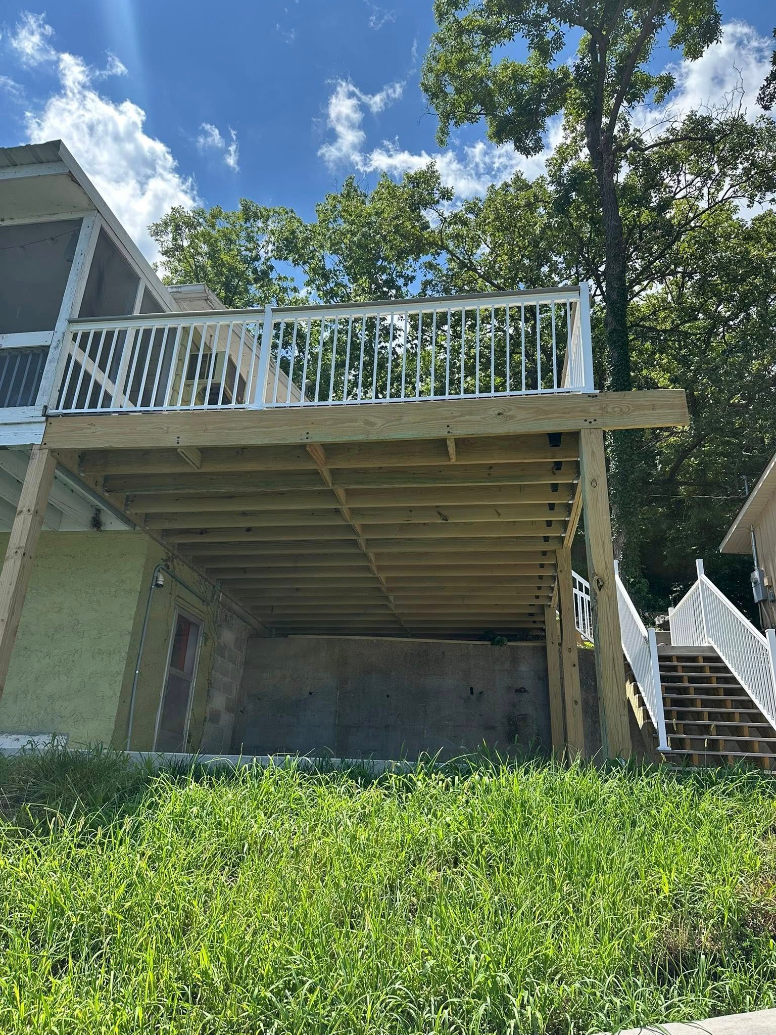 A newly built wooden deck with white railing, supported by posts, overlooking greenery under a blue sky.