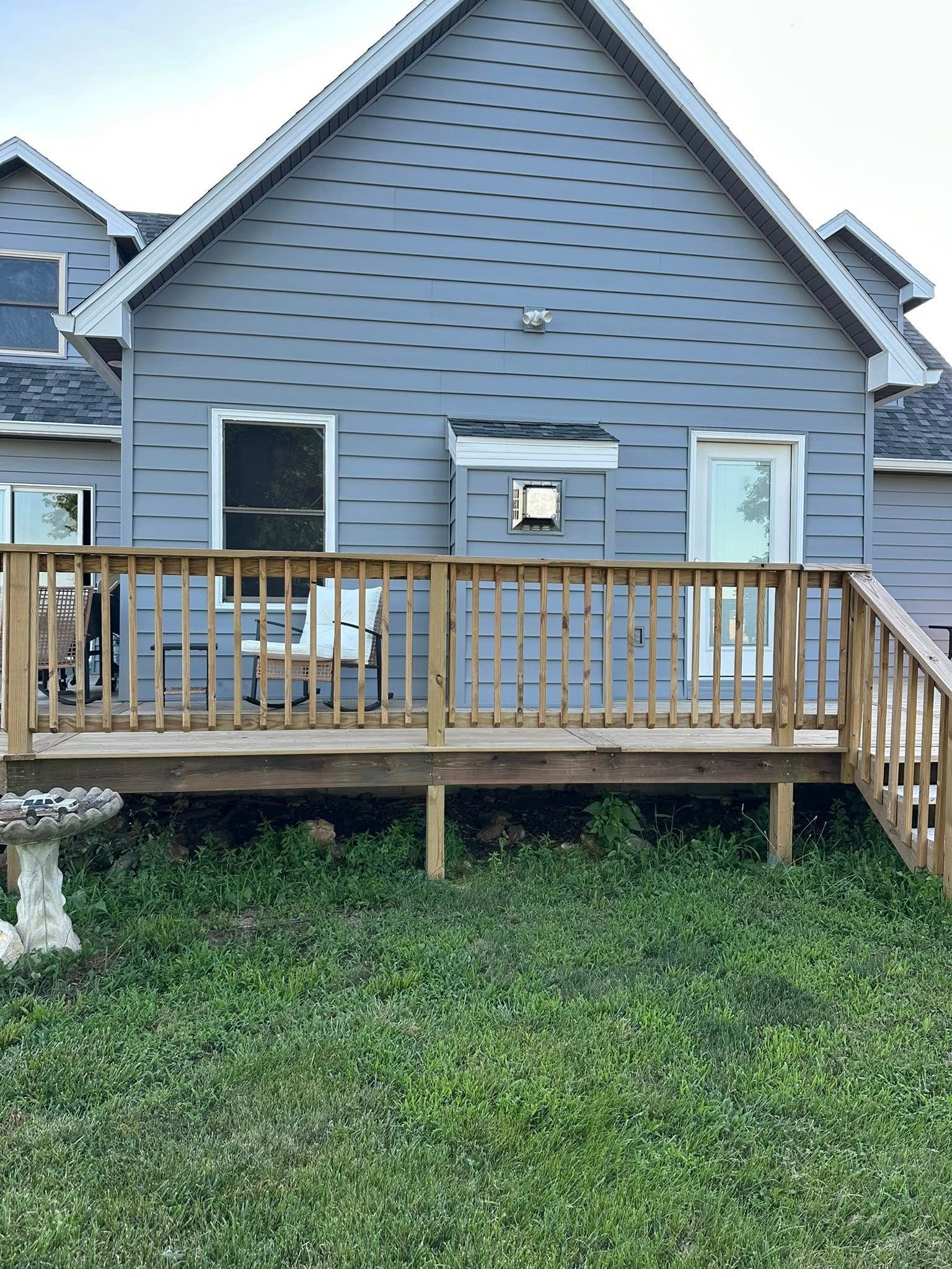 Back of a gray house with a wooden deck and overgrown grass in a yard.