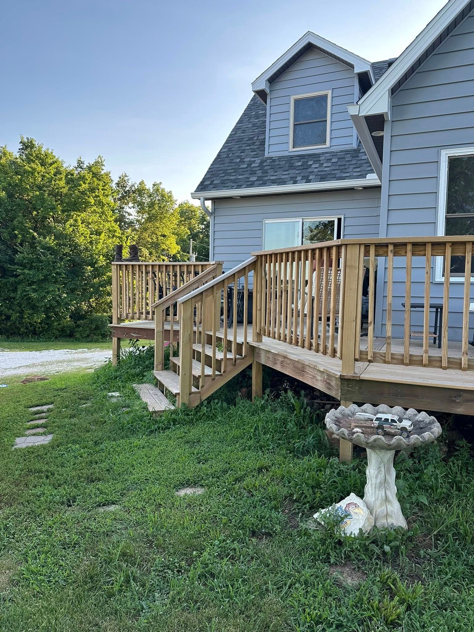 Wooden deck and staircase attached to a gray house, birdbath in the foreground, trees in the background.