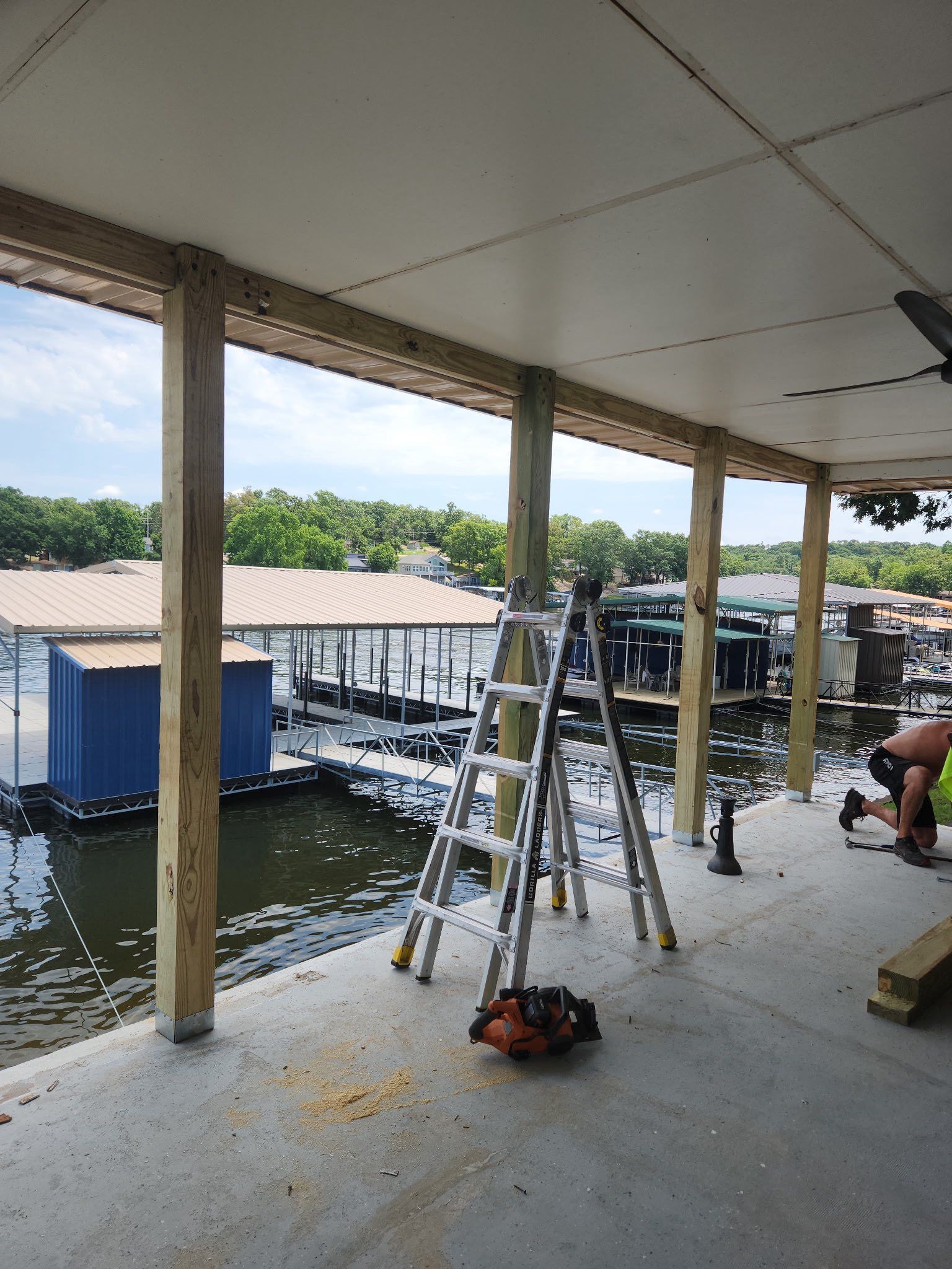 Construction on a waterfront deck; wooden posts, ladders, and tools visible. Someone works nearby.