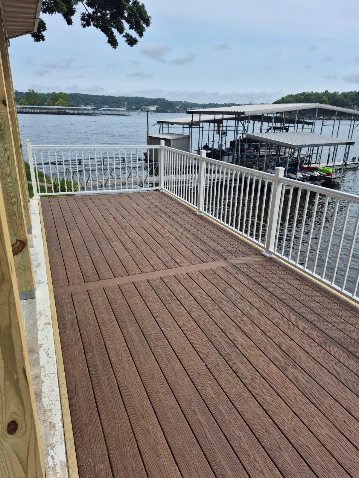 Brown deck overlooking a body of water, white railing. Docks and cloudy sky in the background.