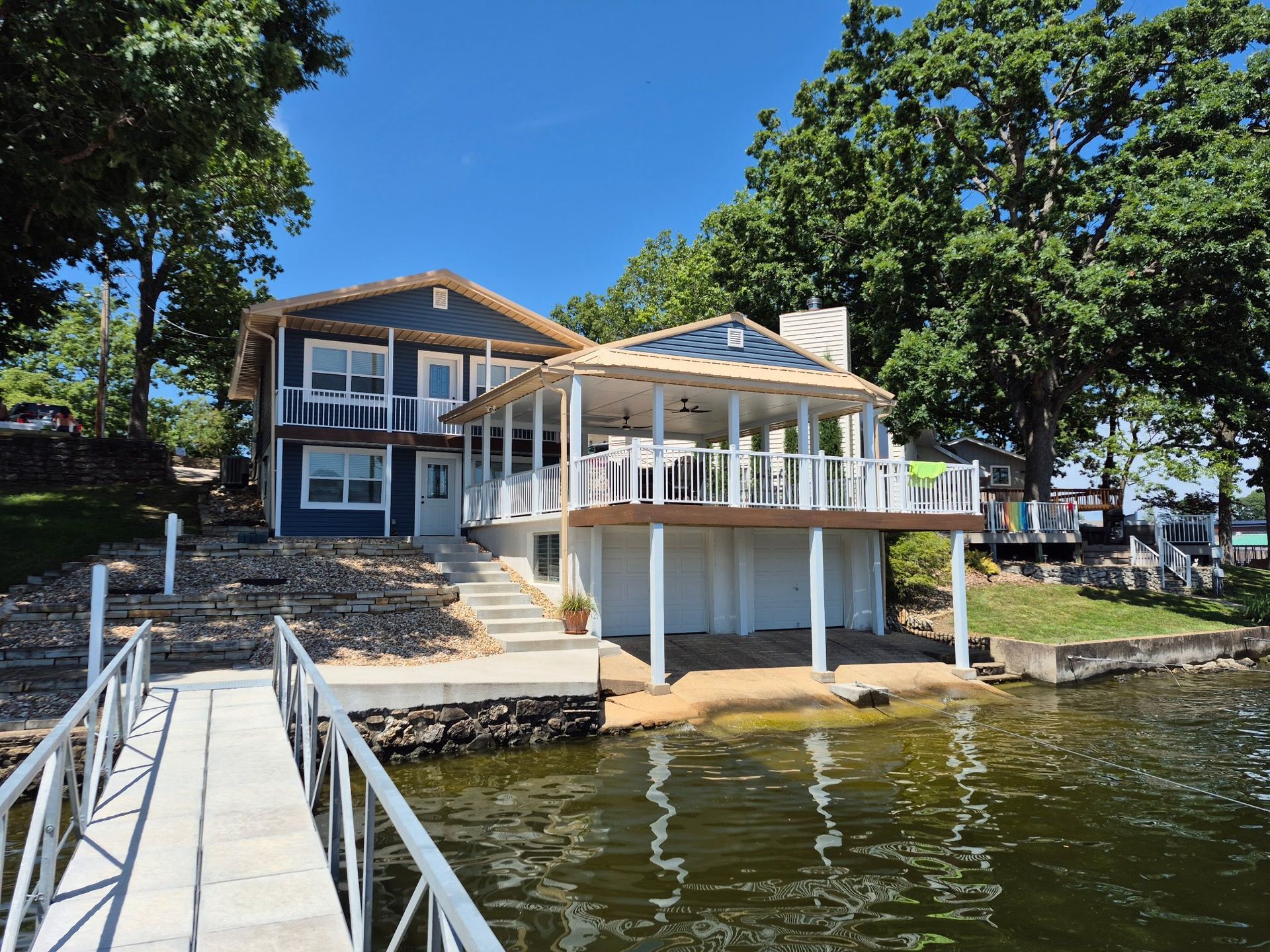 Two-story blue house on a lake with a dock, trees, and a sunny sky.