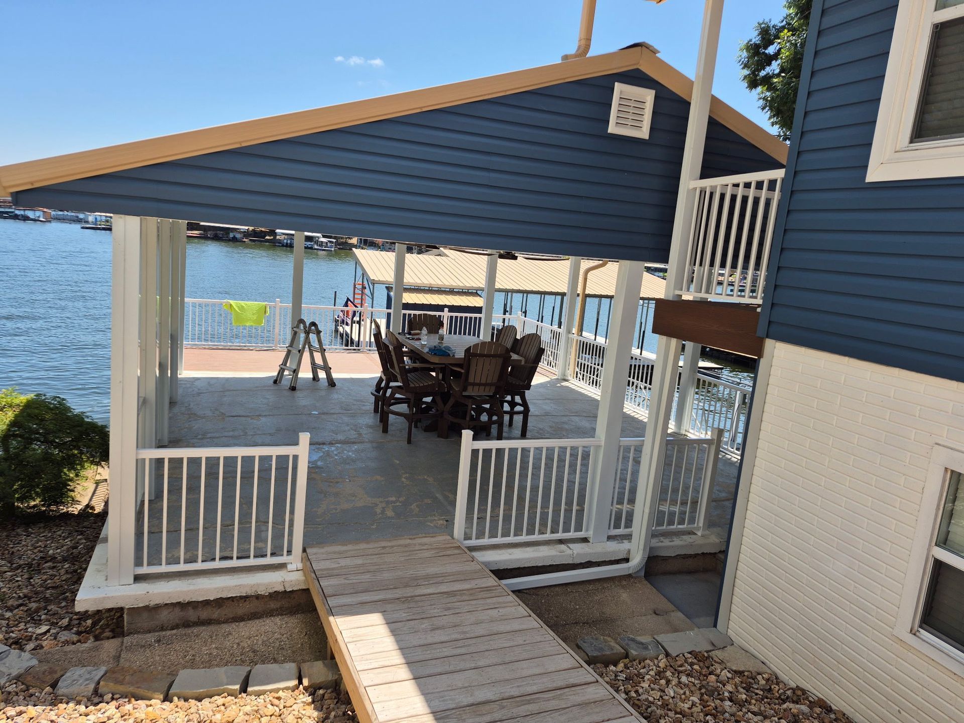 A lakeside deck with a dining area under a blue and tan awning. Wooden walkway leads to the deck.