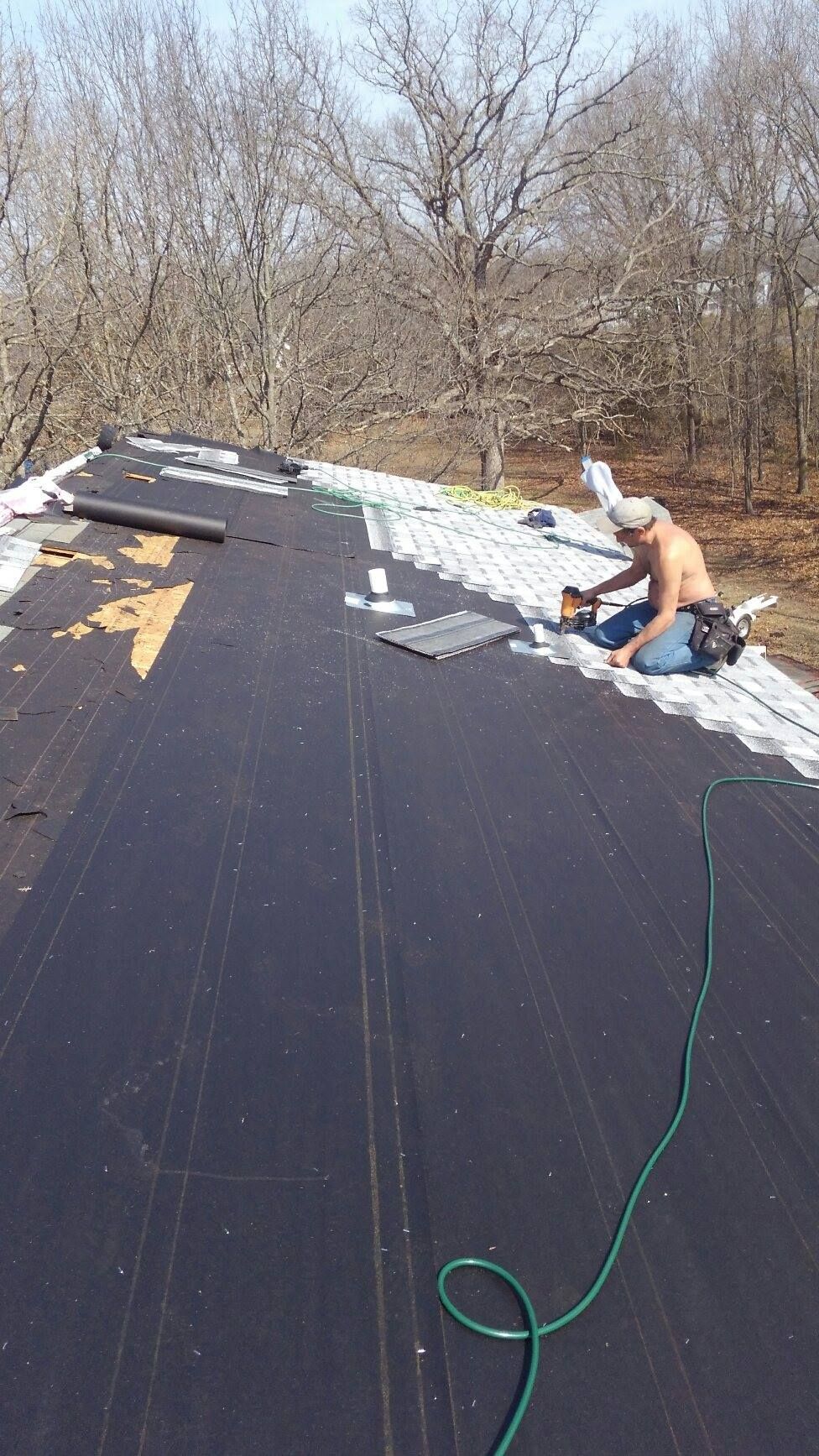 Man working shirtless on a roof, installing metal roofing. Outdoors, trees in background, sunny day.