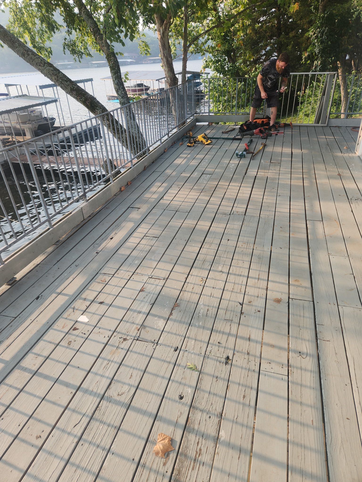 Person on a wooden deck by water, holding tools. Railing and trees are visible. Sunny day.