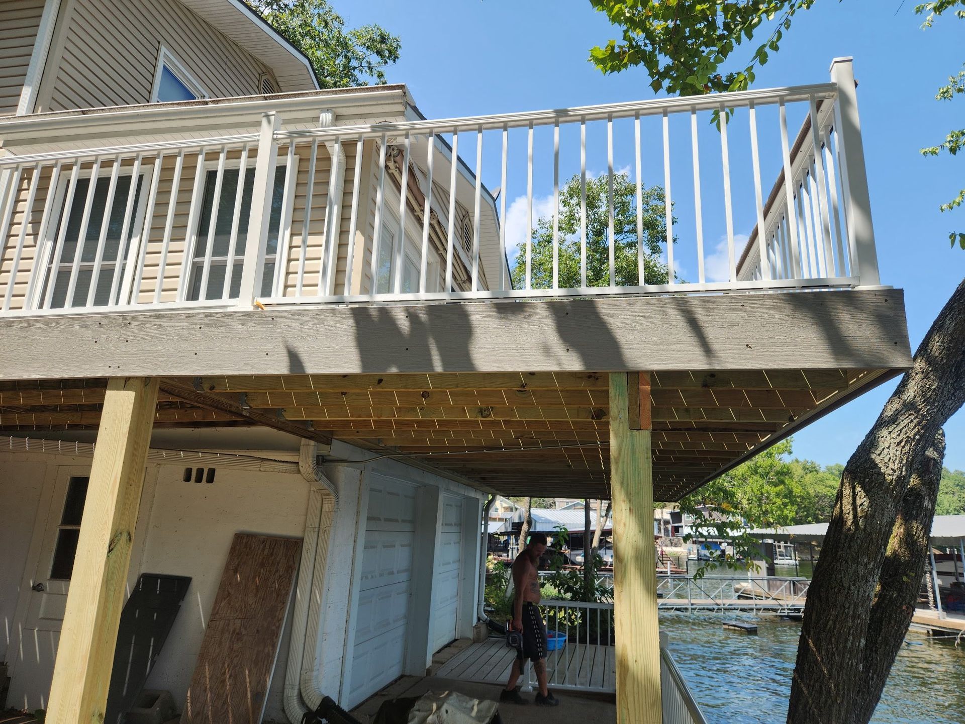 A deck with white railing overhangs a lakeside home. A person stands on the deck below.