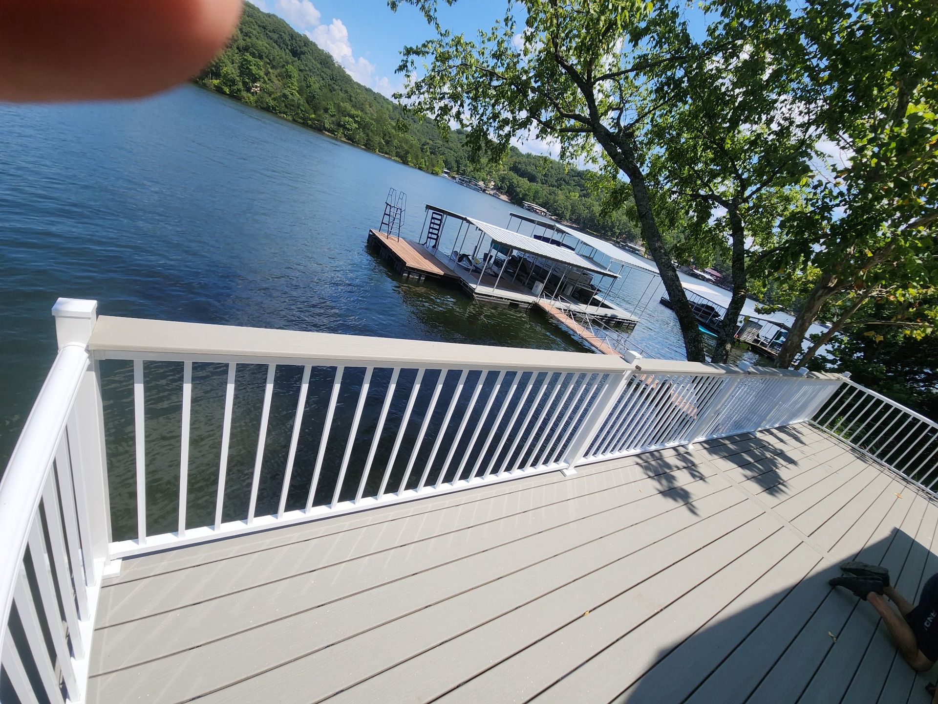 Deck with white railing overlooking a lake with a dock; trees and mountains in the background on a sunny day.