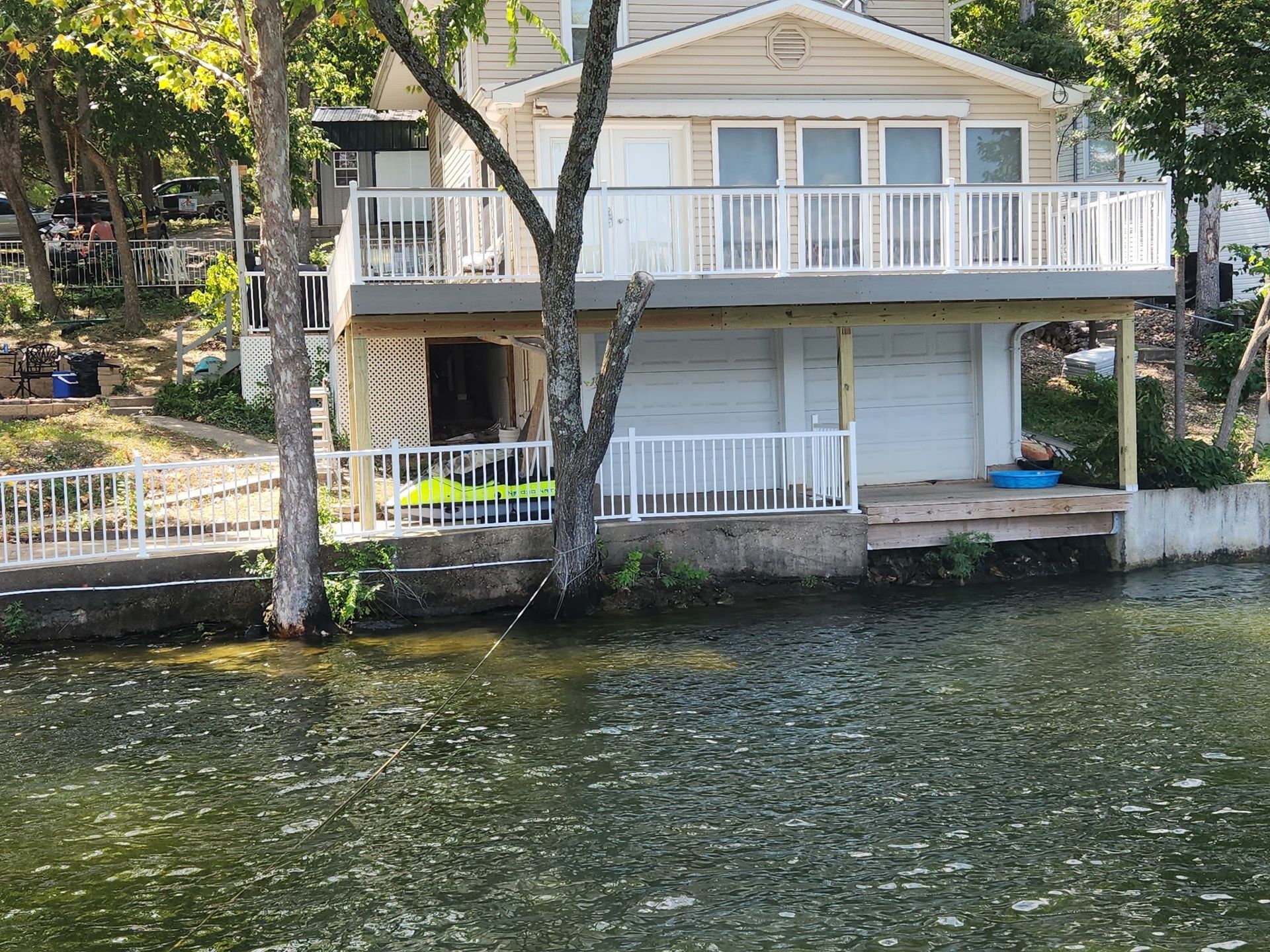 Two-story house with a white railing and deck, on the edge of a lake. Trees stand in front of the house.