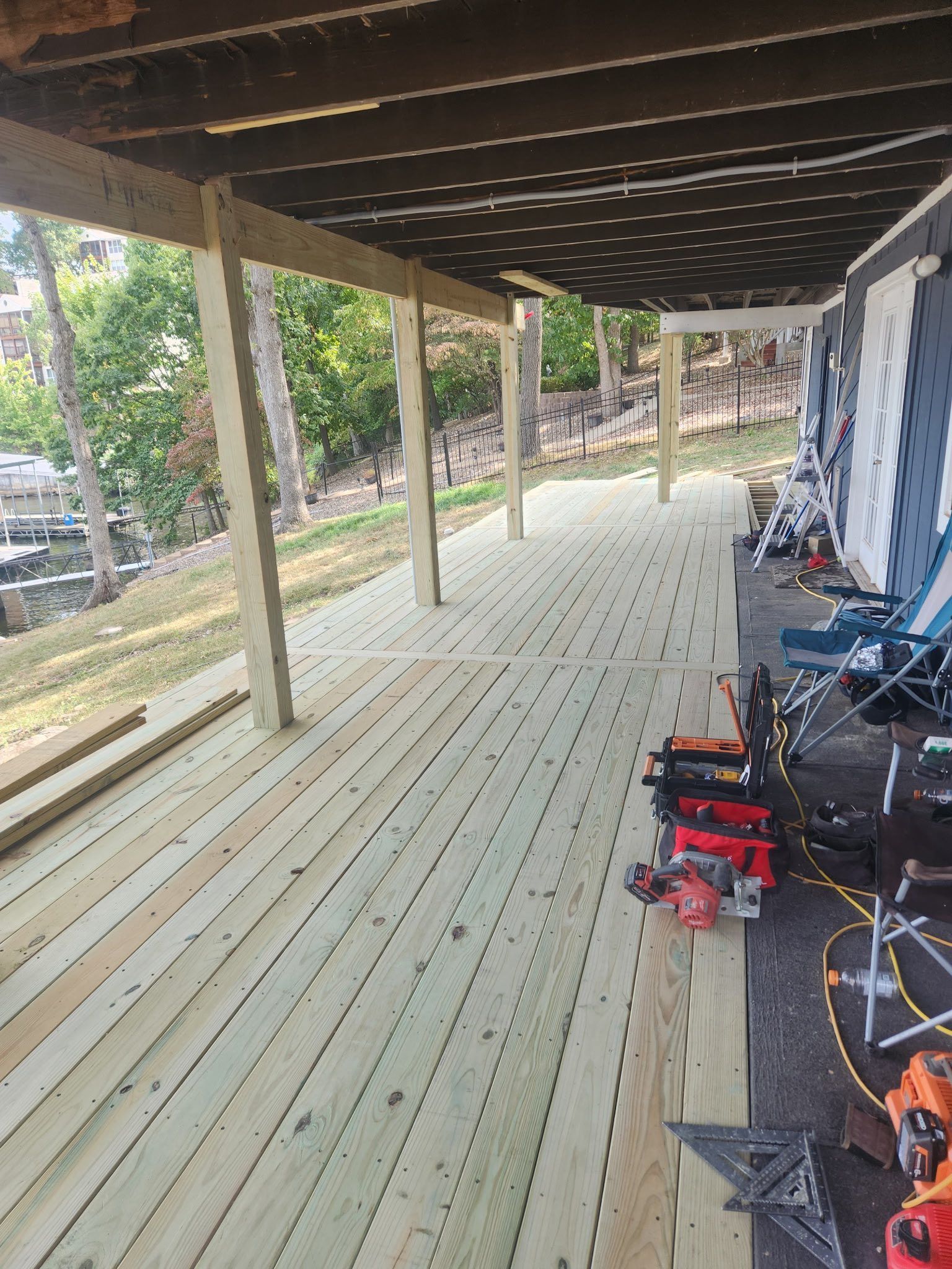 Wooden deck under construction with tools scattered. Supports under a dark-paneled overhang, outdoors with greenery.